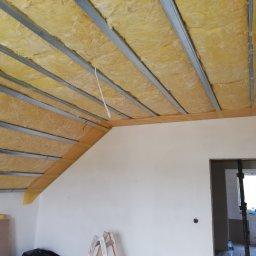 Prace Wykończeniowe - Poddasze w trakcie ocieplania, widoczne metalowe profile sufitowe wypełnione wełną mineralną, fragment ściany z widocznym otworem na gniazdko, drabina w rogu pomieszczenia.
