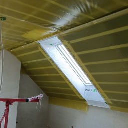 Prace Wykończeniowe - Poddasze w trakcie prac izolacyjnych, widoczne membrany paroizolacyjne w kolorze żółtym, okno dachowe Fakro w folii ochronnej, fragment rusztowania malarskiego i ściana w stanie surowym.