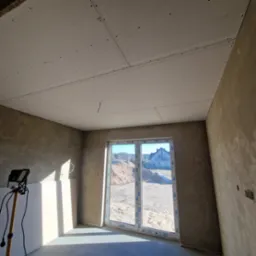 Pokój w trakcie prac wykończeniowych: zamontowana płyta kartonowo-gipsowa na suficie, ściany w surowym tynku, okno z widokiem na plac budowy i dom