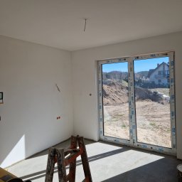 Prace Wykończeniowe - Wnętrze w stanie deweloperskim z oknem balkonowym VEKA, widoczne ślady po gładzi, wystający kabel z sufitu, drabina malarska i szpachla w rogu. Na zewnątrz widok na hałdę piachu i dom.