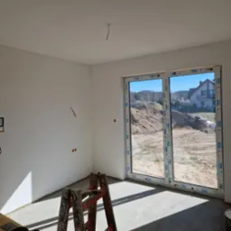 Wnętrze w stanie deweloperskim z oknem balkonowym VEKA, widoczne ślady po gładzi, wystający kabel z sufitu, drabina malarska i szpachla w rogu. Na zewnątrz widok na hałdę piachu i dom.