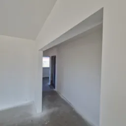 Wnętrze świeżo pomalowanego pokoju z widocznym otworem drzwiowym bez drzwi i oknem w tle, surowy beton na podłodze, białe ściany i sufit.