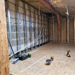 Prace Wykończeniowe - Montaż metalowego stelażu do płyty karton gips na ścianie z pustaków, widoczne narzędzia: poziomica laserowa, szlifierka kątowa oraz wkrętarka, podłoga wyłożona płytami OSB.