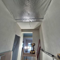 Prace Wykończeniowe - Montaż izolacji termicznej sufitu folią aluminiową w Pogórzu. Widoczna drabina, wkrętarka i poziomica. Prace wykończeniowe w trakcie realizacji.