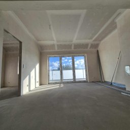 Prace Wykończeniowe - Surowe wnętrze z oknem balkonowym, ściany przygotowane pod malowanie, widoczne płyty gipsowe na suficie i profile metalowe. Jasne, przestronne pomieszczenie w trakcie prac wykończeniowych.