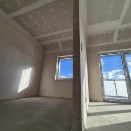 Prace Wykończeniowe „Zuchy” - Wnętrze w trakcie wykończenia: surowy beton na podłodze, ściany po szpachlowaniu, płyty karton-gips na suficie z widocznymi punktami szpachlowania. Okno i drzwi balkonowe z PCV.
