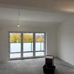 Prace Wykończeniowe - Białe, świeżo pomalowane wnętrze z dużym oknem balkonowym i widokiem na jesienne drzewa. Na podłodze stoi urządzenie budowlane. Minimalistyczny styl wykończenia.