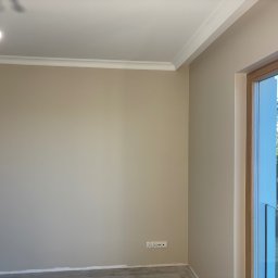 Prace Wykończeniowe - Beżowa ściana po malowaniu z białym sztukateryjnym wykończeniem sufitu i drewnianą ramą okienną. Widoczne gniazdka elektryczne i żarówka.