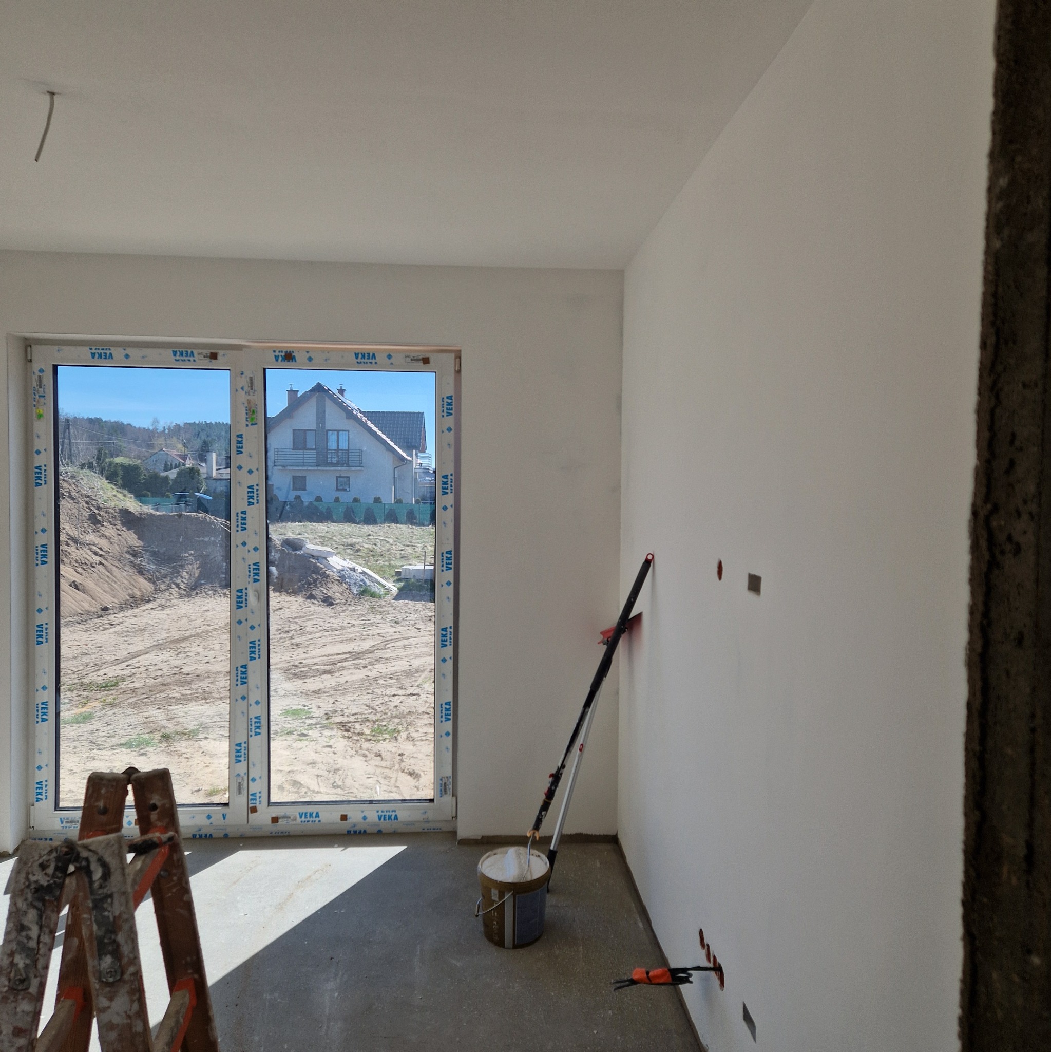 Wnętrze w trakcie prac wykończeniowych: pomalowana na biało ściana z widocznymi otworami na gniazdka elektryczne, drabina malarska i wiadro z farbą przy ścianie, okno z widokiem na budowę i dom w tle.