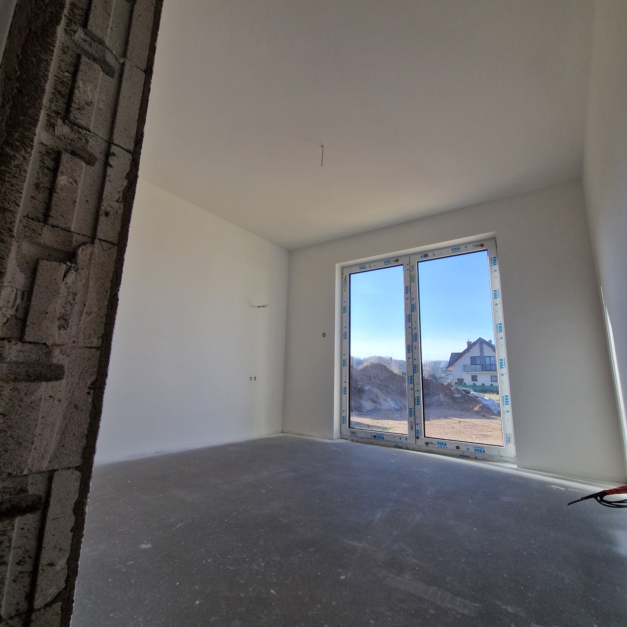 Surowe wnętrze pokoju z widokiem na dom przez okno, ściany pomalowane na biało, surowy beton na podłodze, widoczny fragment nieotynkowanego muru.