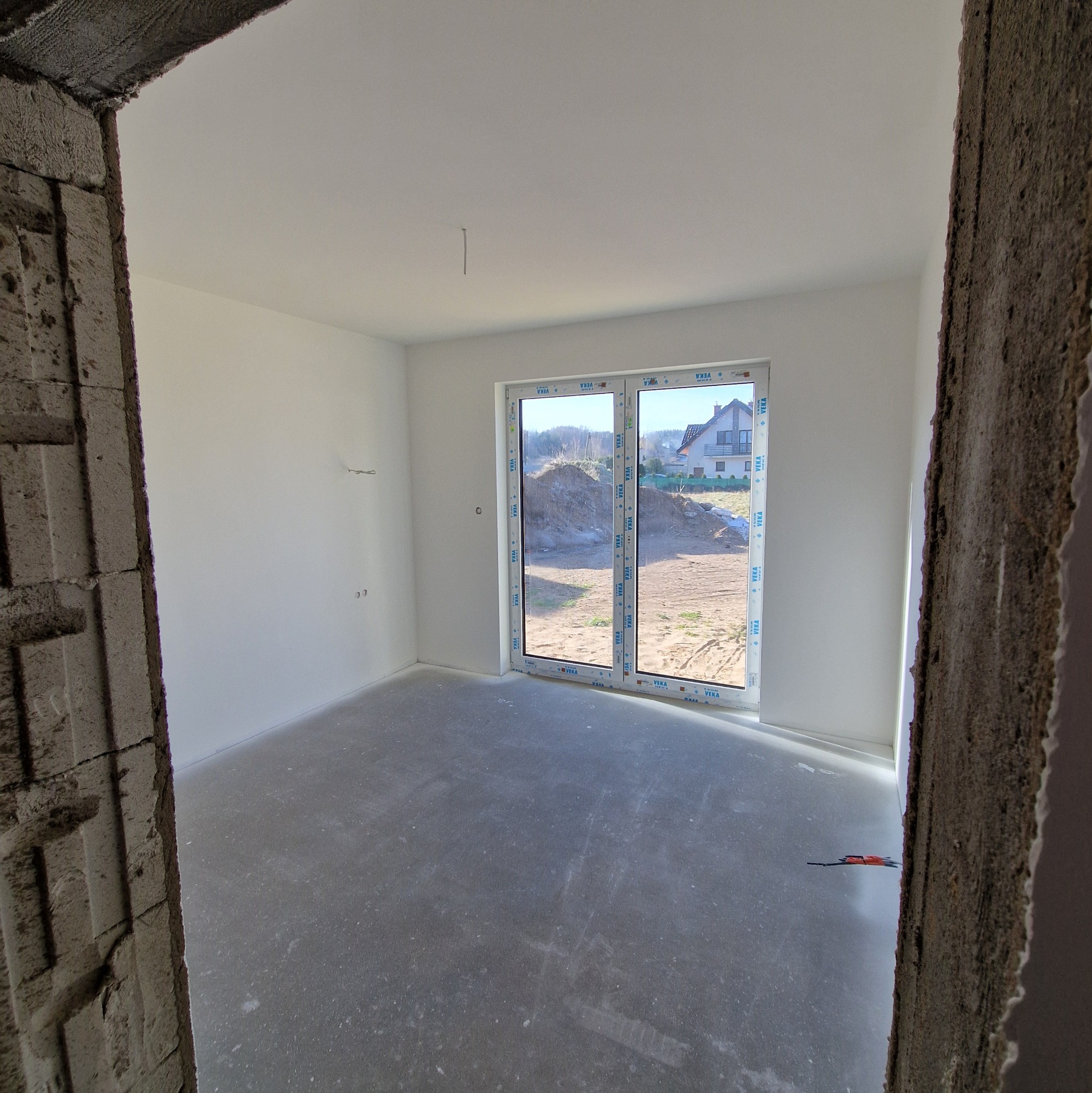 Surowe wnętrze domu w trakcie remontu, widoczne gładkie, białe ściany, betonowa podłoga, okno balkonowe z widokiem na ogród i niedokończone obramowanie drzwi z surowego betonu.