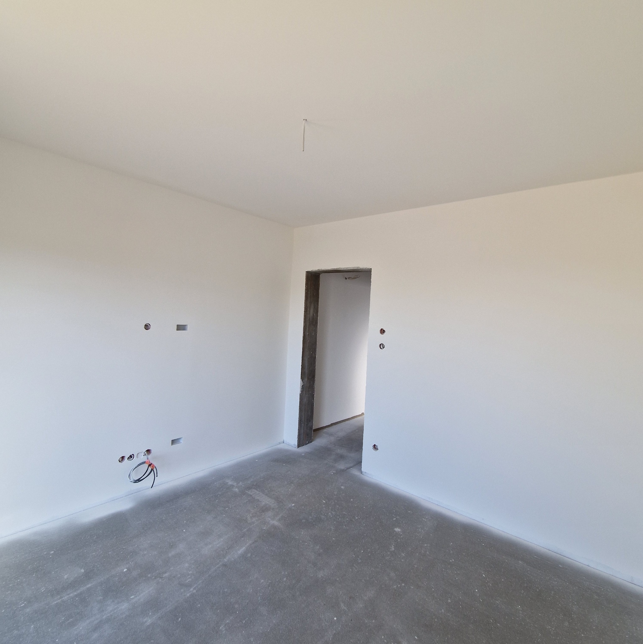 Wnętrze w stanie deweloperskim z białymi ścianami, surowym betonowym podłożem, widocznymi puszkami elektrycznymi i otwartym przejściem do innego pomieszczenia.