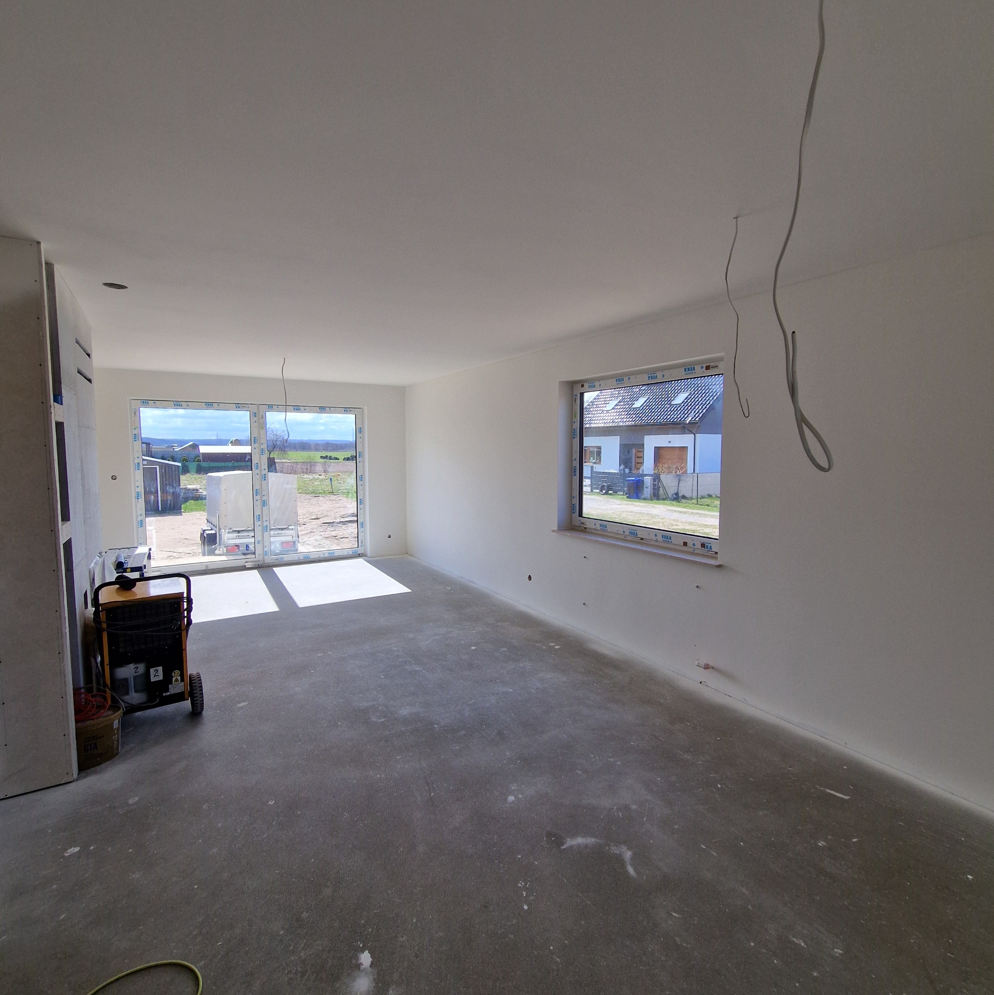 Wnętrze w stanie deweloperskim w Pogórzu po szpachlowaniu, z widocznymi przewodami zwisającymi z sufitu, grzejnikiem budowlanym i ścianami przygotowanymi pod malowanie.