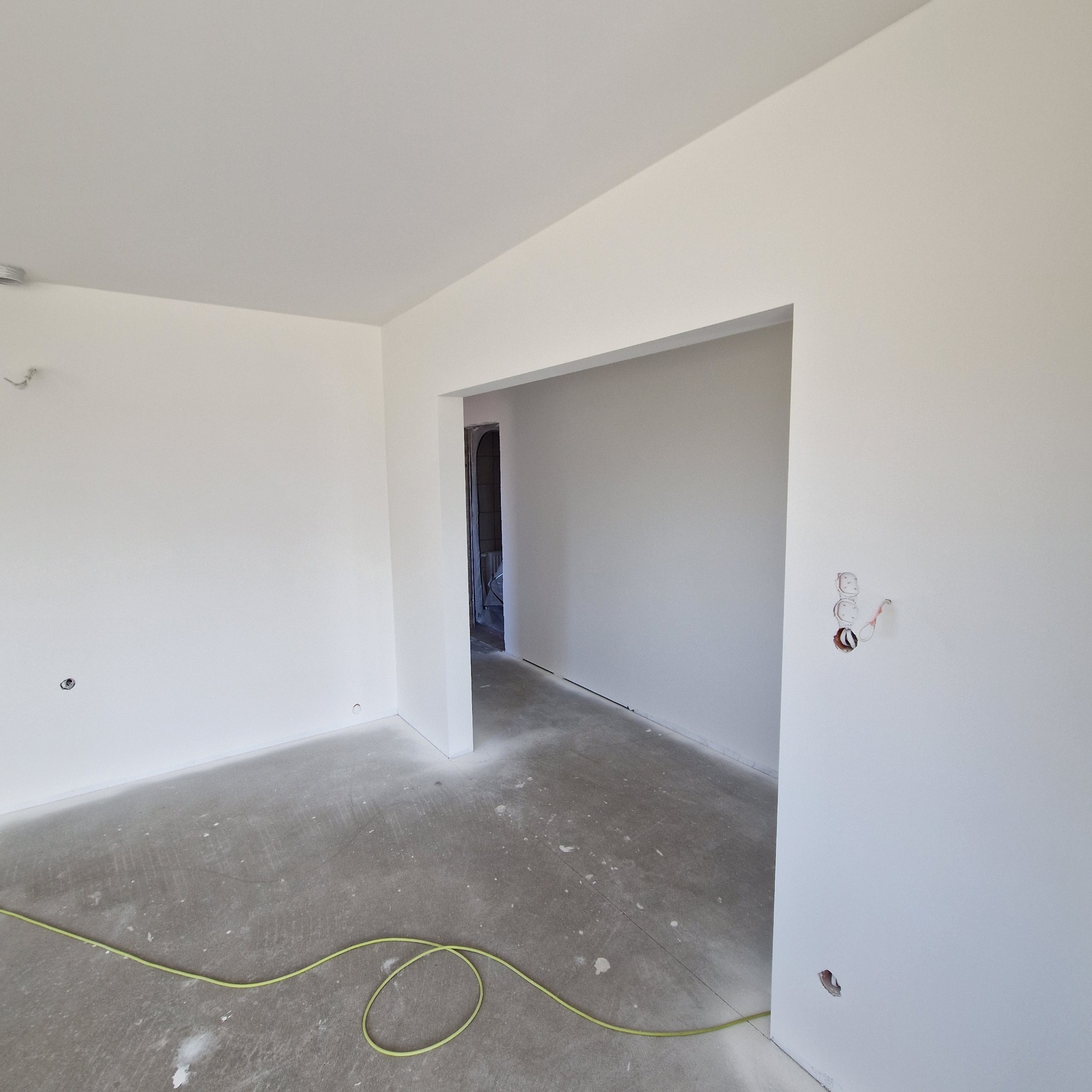 Wnętrze pokoju po białym malowaniu ścian i sufitu, widoczne gniazdka elektryczne bez osprzętu, kabel na podłodze i surowy beton. Otwór drzwiowy prowadzi do kolejnego pomieszczenia.