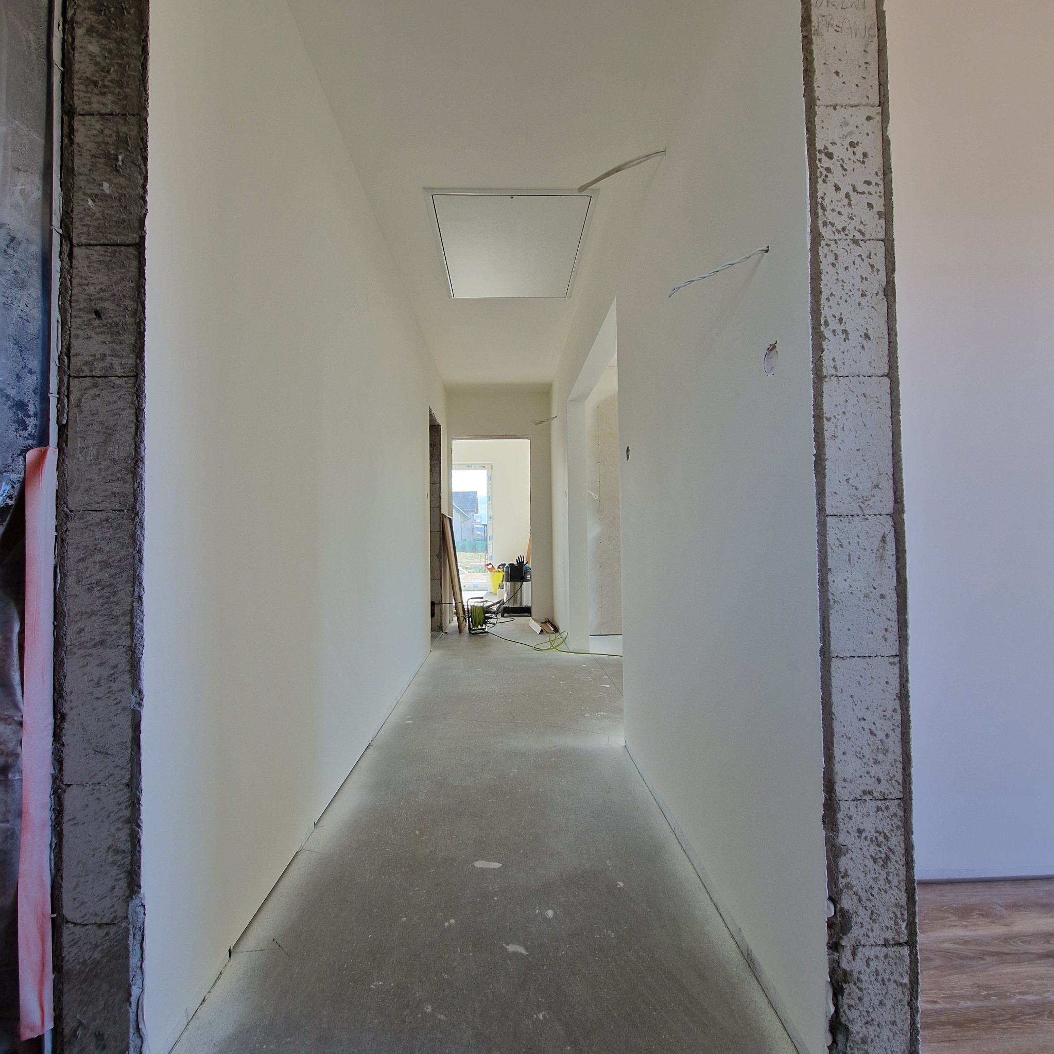 Korytarz w trakcie prac wykończeniowych, ściany pokryte gładzią, surowy beton na podłodze, widoczne narzędzia i akcesoria w głębi pomieszczenia.