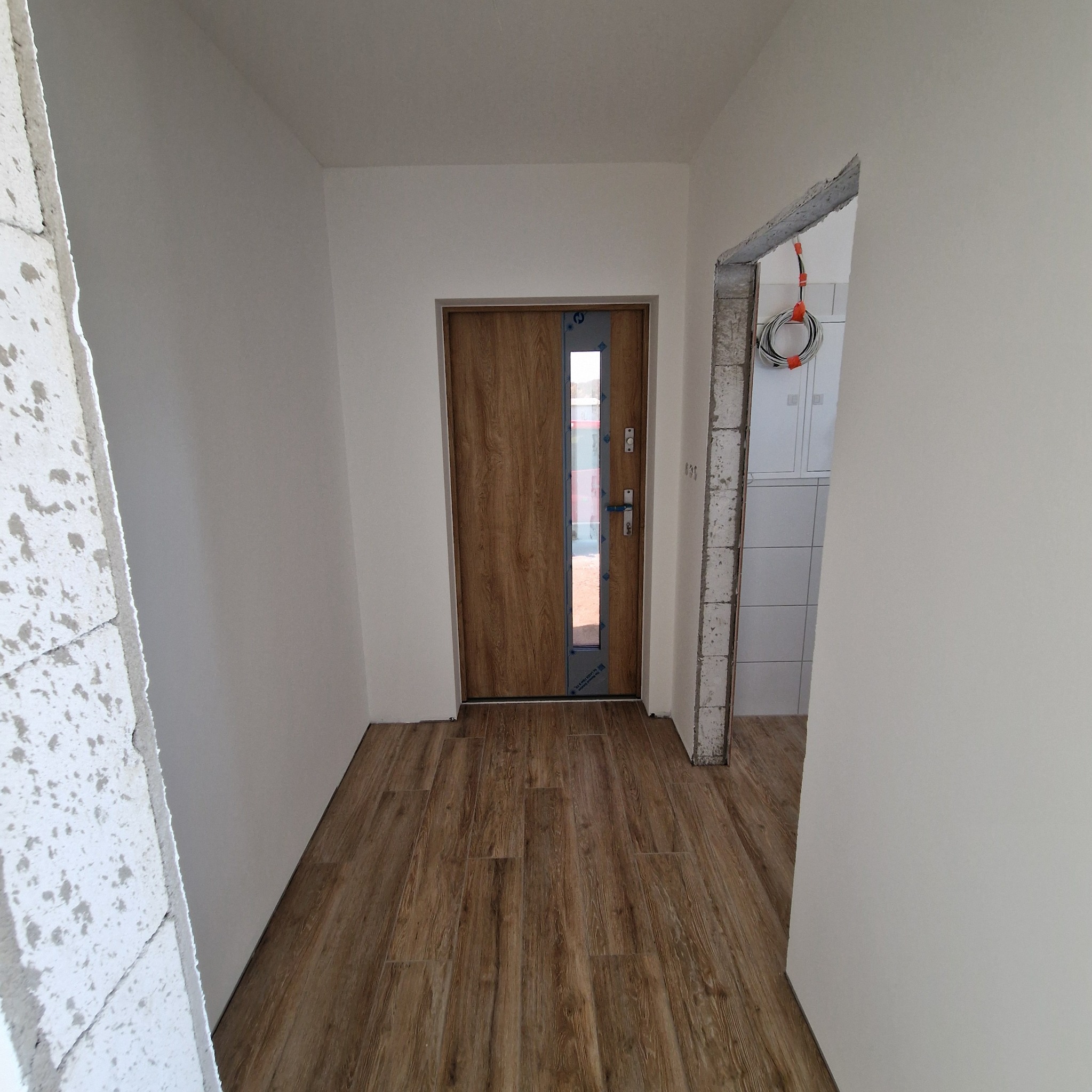 Korytarz w trakcie remontu z nową drewnopodobną podłogą, zamontowanymi drzwiami z przeszkleniem i surowymi ścianami w tle