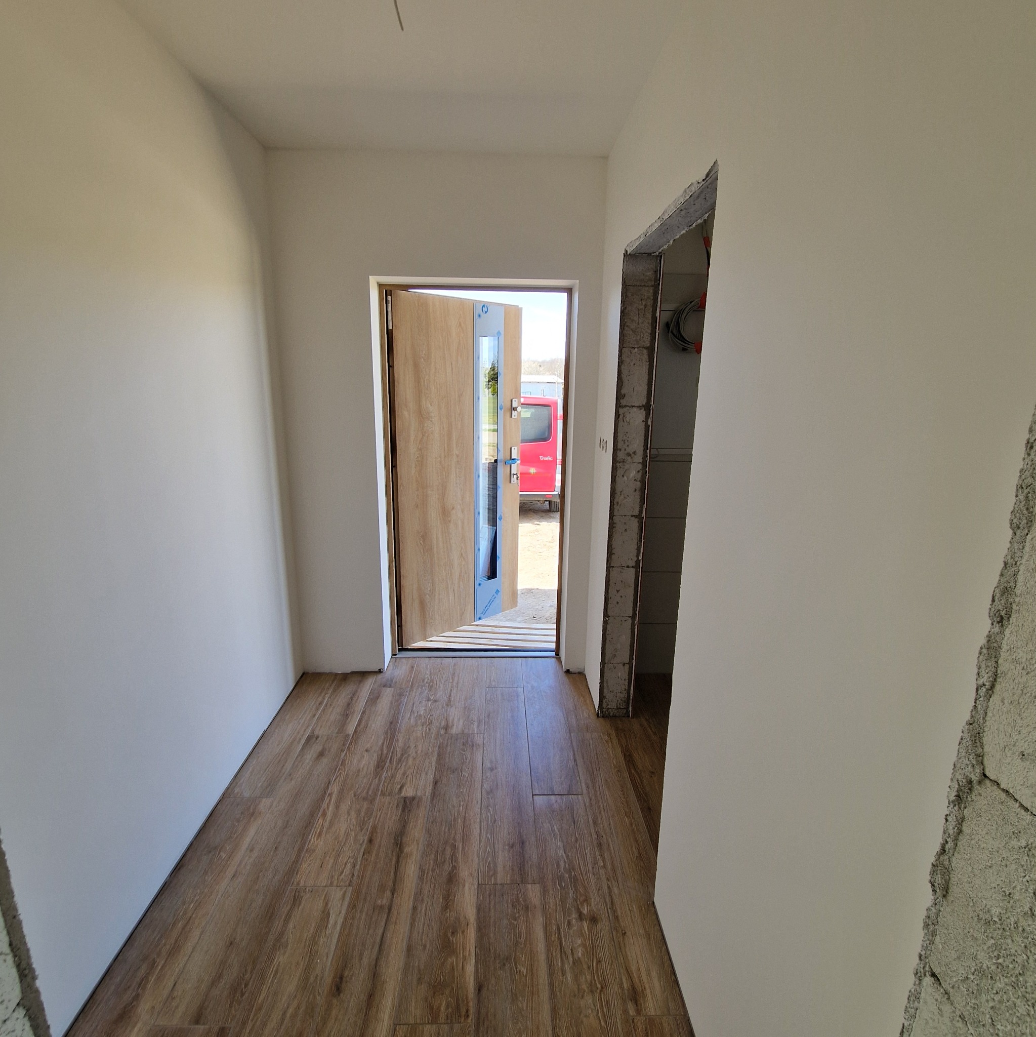 Wnętrze w trakcie remontu: świeżo ułożone panele podłogowe w odcieniach brązu, białe ściany po malowaniu, częściowo zamontowane drzwi z przeszkleniem i surowy otwór drzwiowy z widocznymi przewodami.