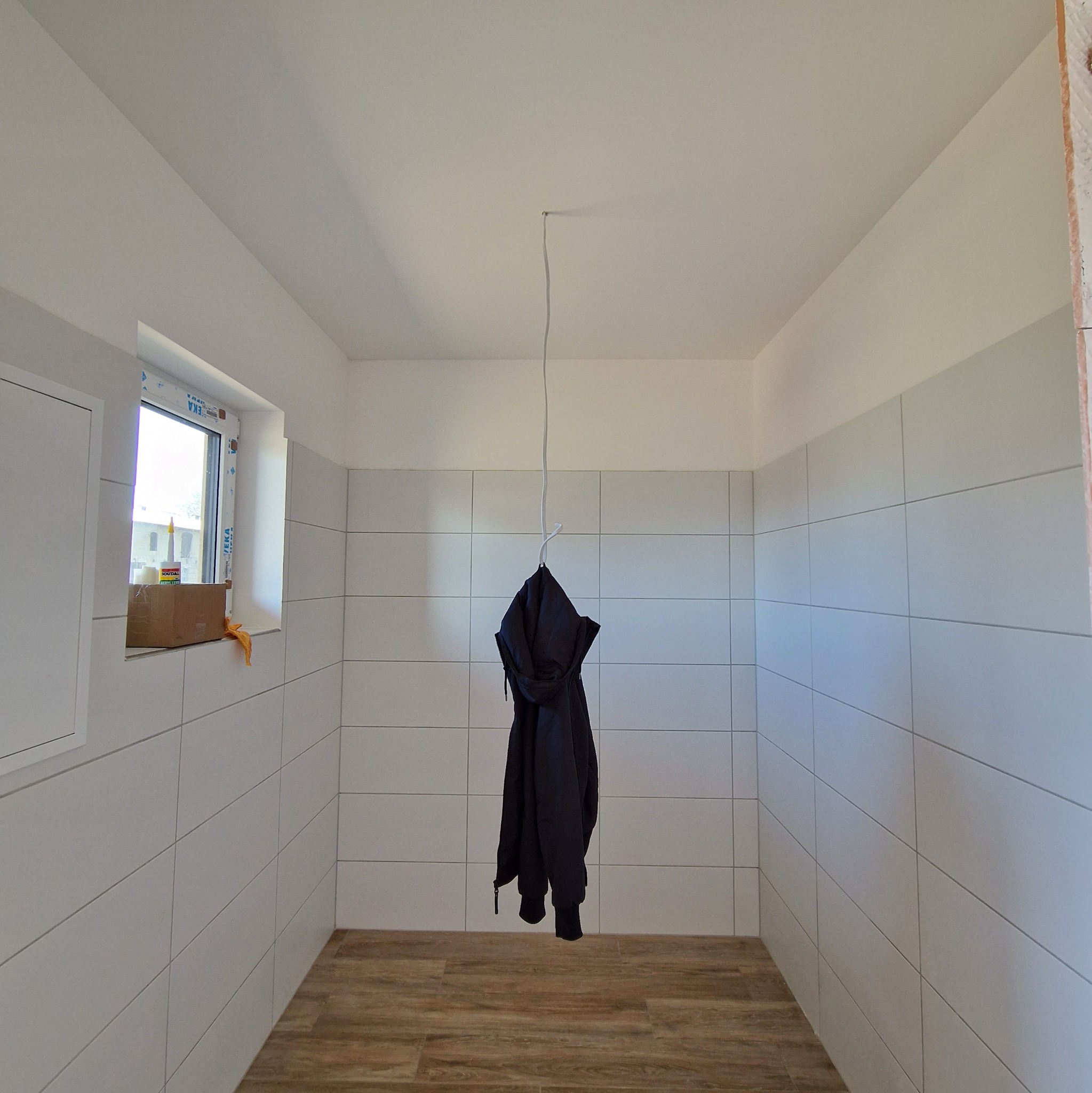 Wnętrze w trakcie prac wykończeniowych, ściany wyłożone białymi płytkami, na podłodze panele, kurtka zawieszona na haku pod sufitem, w oknie karton z materiałami budowlanymi.