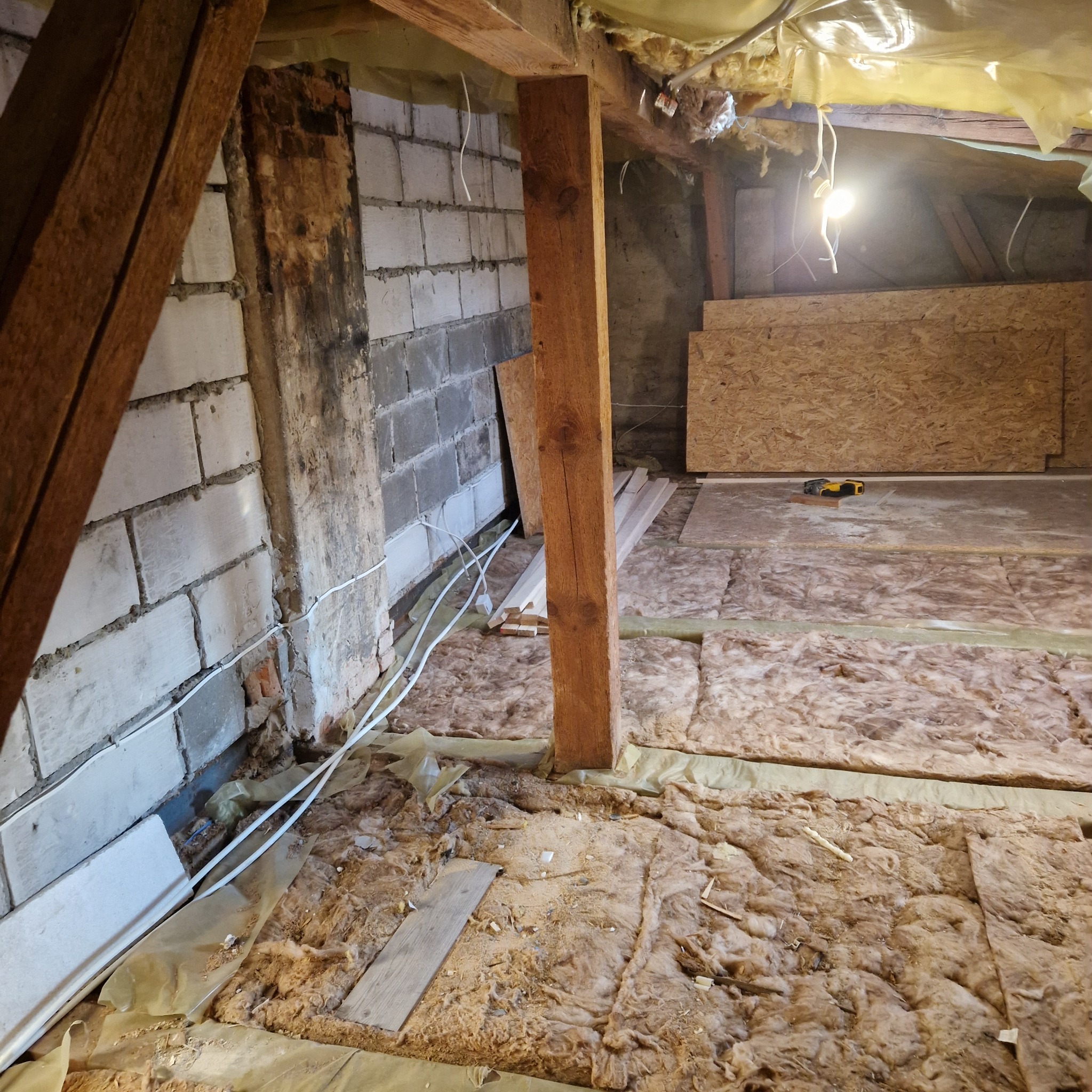 Adaptacja poddasza: izolacja wełną mineralną, fragment ściany z bloczków, drewniane belki konstrukcyjne, widoczne przewody elektryczne i płyty OSB.