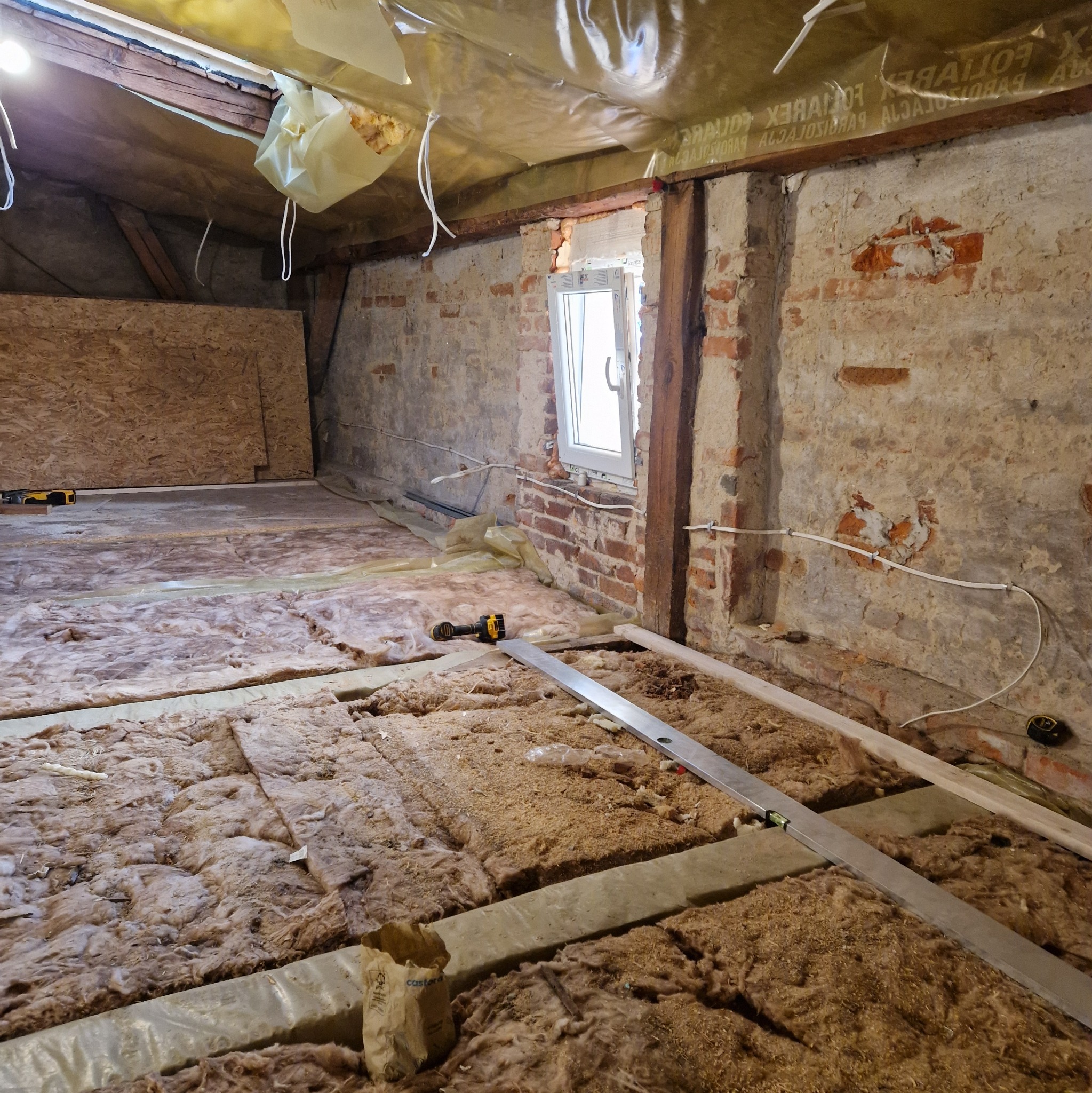 Poddasze w trakcie ocieplania: wełna mineralna między legarami, poziomica, w tle odsłonięta cegła i okno.