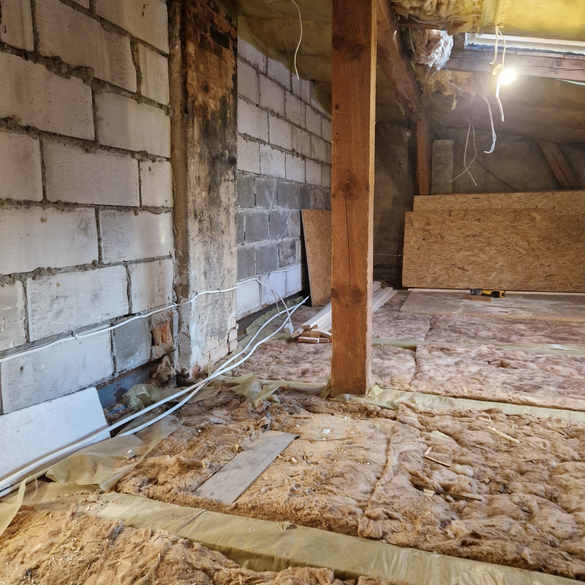 Poddasze w trakcie ocieplania: wełna mineralna rozłożona na podłodze, widoczne przewody elektryczne, drewniane belki konstrukcyjne i fragment ściany z bloczków betonowych.
