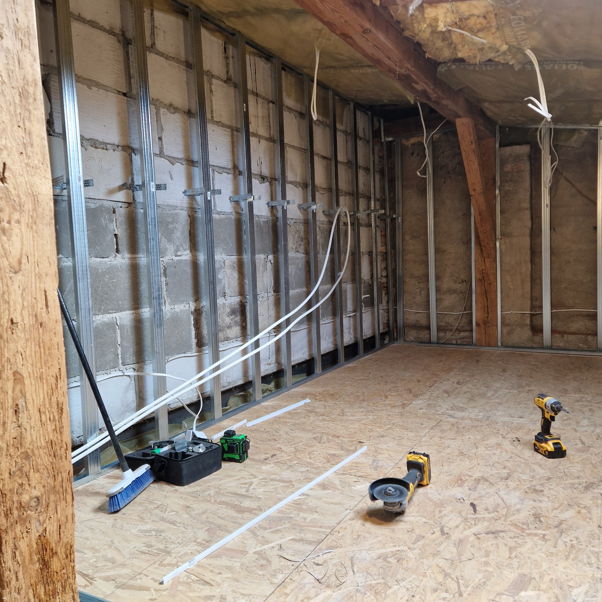 Montaż metalowego stelażu do płyty karton gips na ścianie z pustaków, widoczne narzędzia: poziomica laserowa, szlifierka kątowa oraz wkrętarka, podłoga wyłożona płytami OSB.