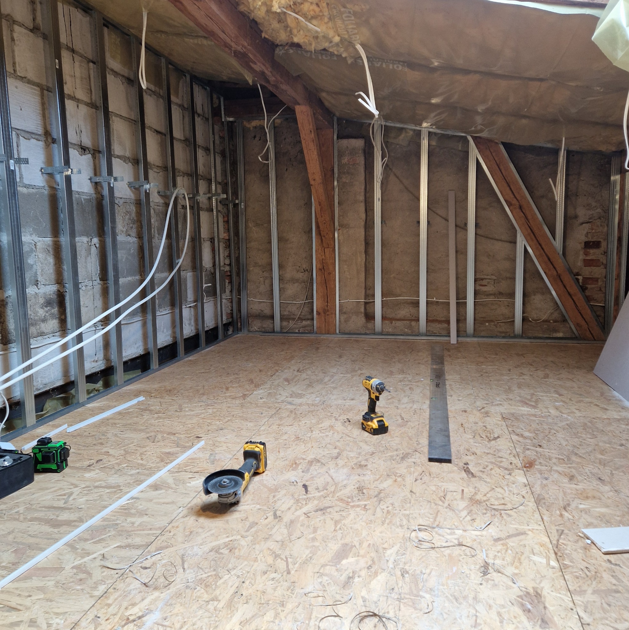 Adaptacja poddasza: konstrukcja szkieletowa z profili stalowych, izolacja stropu, płyta OSB na podłodze, narzędzia (szlifierka kątowa, wkrętarka) i poziomica laserowa na posadzce.