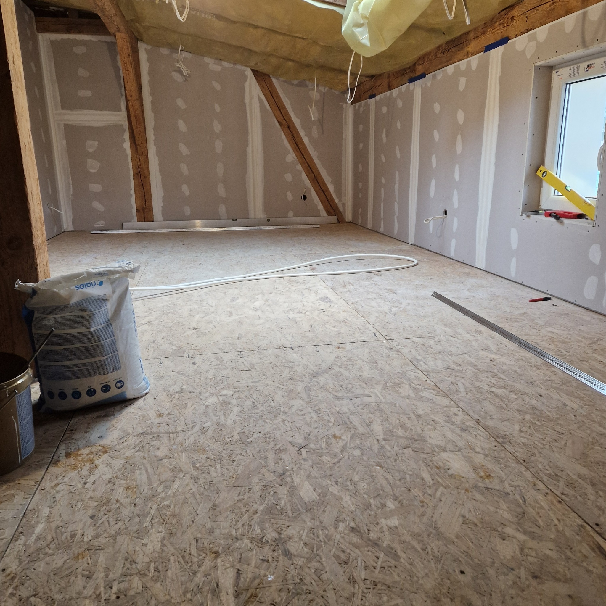 Poddasze w trakcie adaptacji: ściany pokryte płytami kartonowo-gipsowymi, widoczne łączenia wymagające wykończenia, podłoga z płyt OSB, worek zaprawy i poziomica przy oknie.