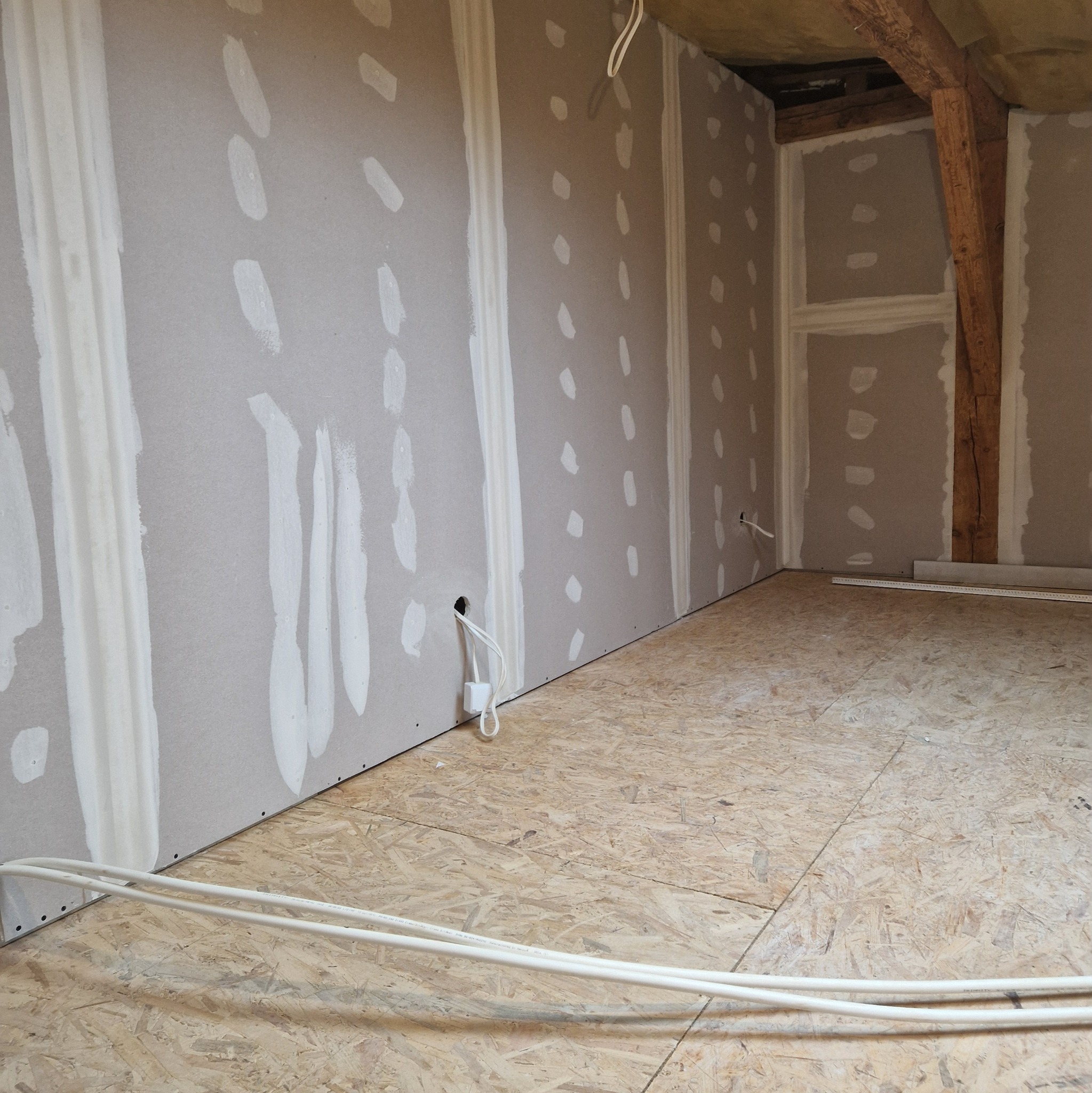 Wnętrze w trakcie prac wykończeniowych: ściany z płyt kartonowo-gipsowych z widocznymi łączeniami wypełnionymi masą, podłoga z płyt OSB, wystające przewody elektryczne oraz fragment drewnianej...