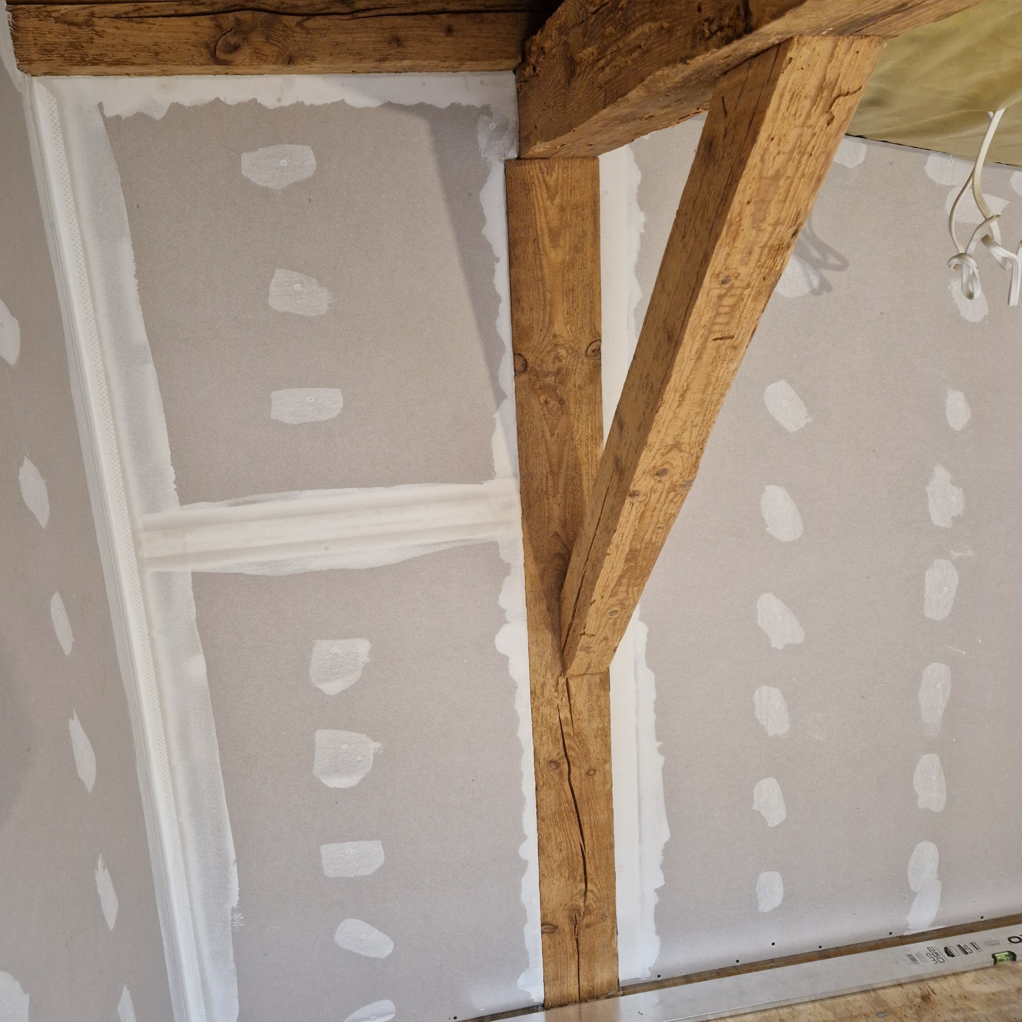 Montaż płyty karton gips na ścianie z drewnianą belką, widoczne łączenia wzmocnione taśmą i częściowo zaszpachlowane.