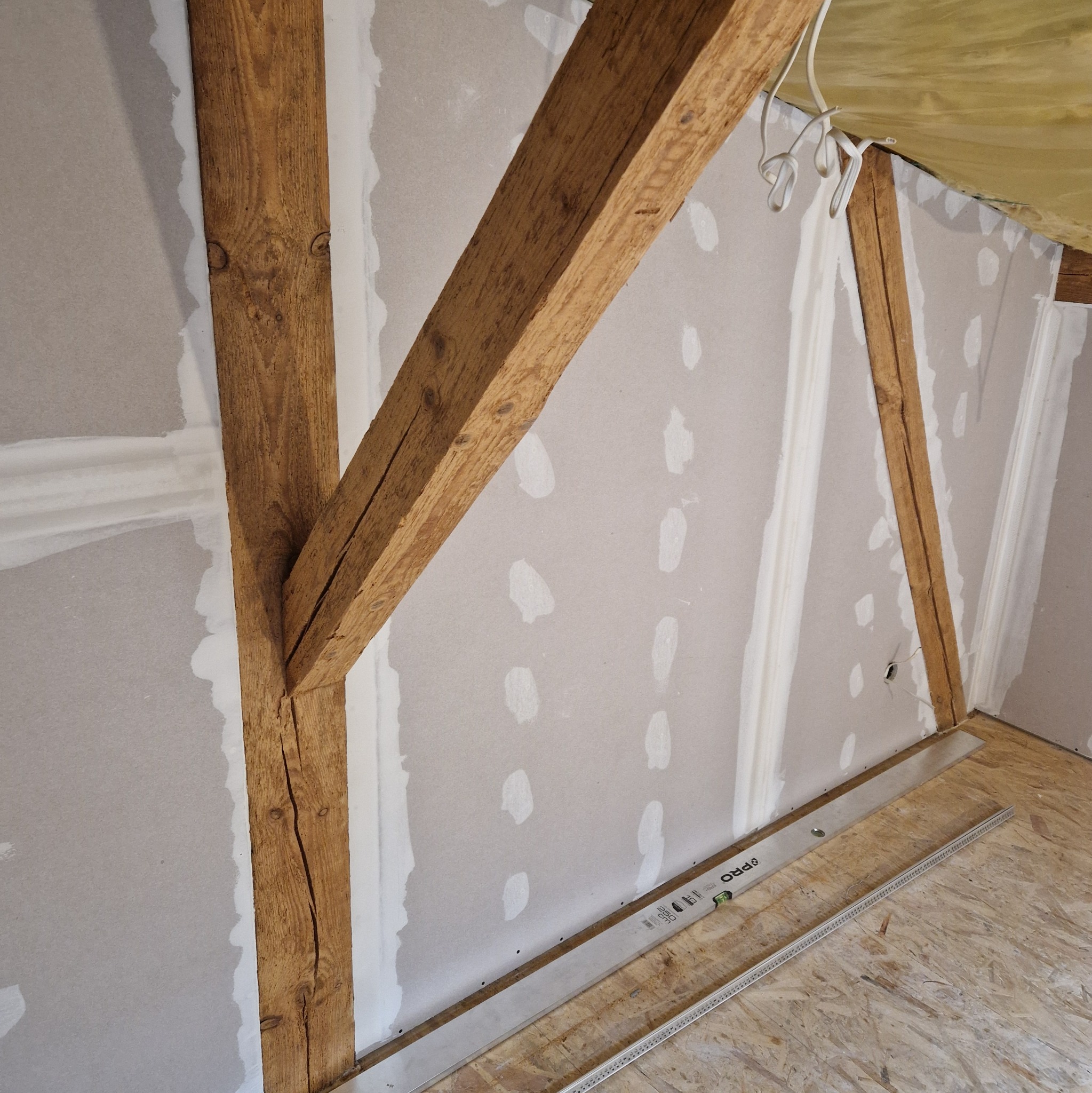 Poddasze w trakcie remontu: widoczne drewniane belki konstrukcyjne, płyty kartonowo-gipsowe z widocznymi miejscami po szpachlowaniu, poziomica na podłodze z płyty OSB.
