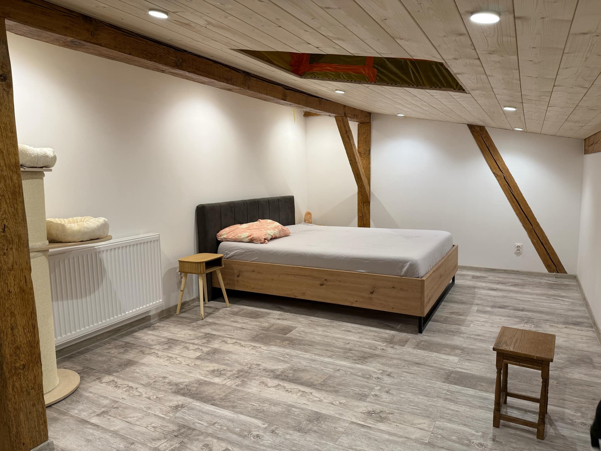 Pokój na poddaszu z drewnianą podłogą imitującą deski, drewnianymi belkami stropowymi, białymi ścianami, łóżkiem, stolikiem nocnym i drapakiem dla kota.