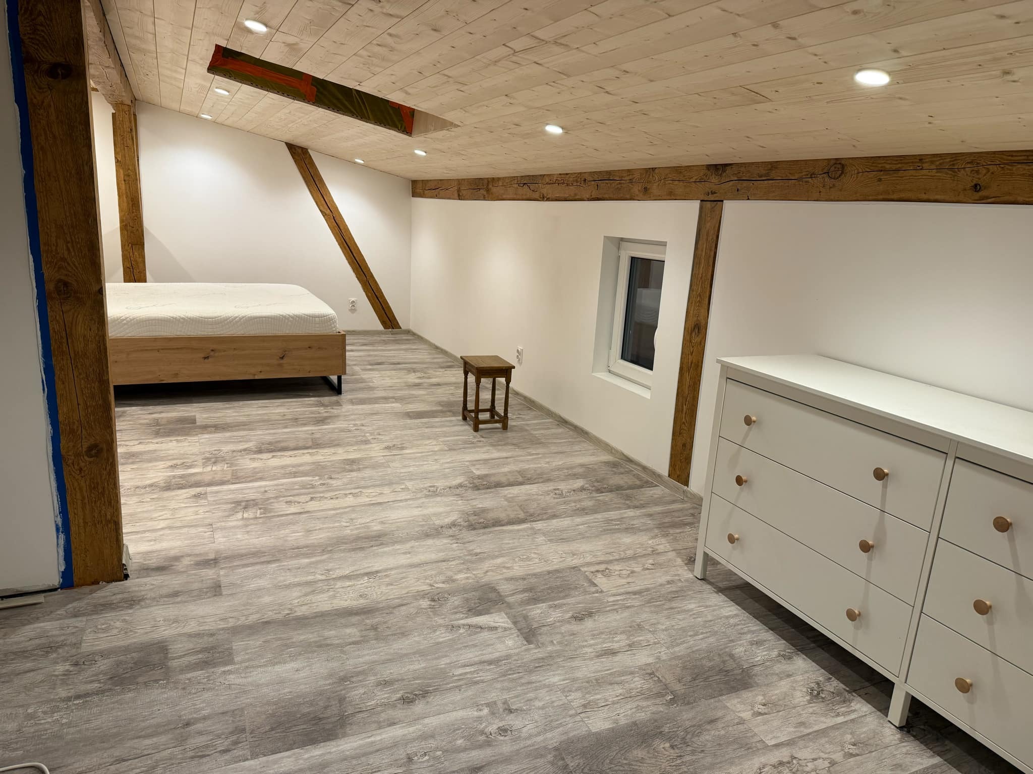 Pokój na poddaszu z drewnianym sufitem, panelami podłogowymi imitującymi drewno, białymi ścianami, łóżkiem, komodą i małym stołkiem.