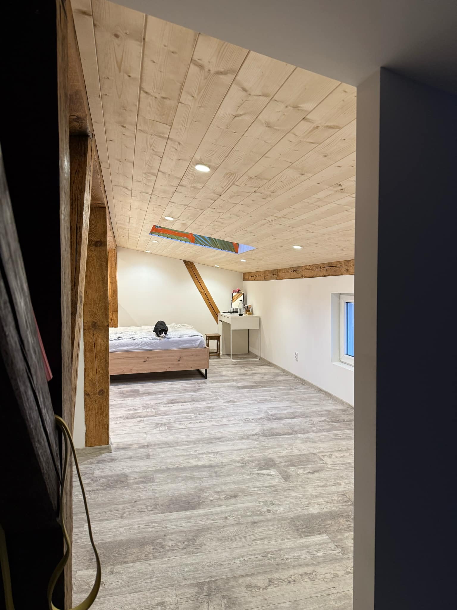 Wykończone poddasze w Pogórzu z drewnianym sufitem, panelami podłogowymi i widocznymi belkami konstrukcyjnymi, częściowo umeblowane z kotem na łóżku.