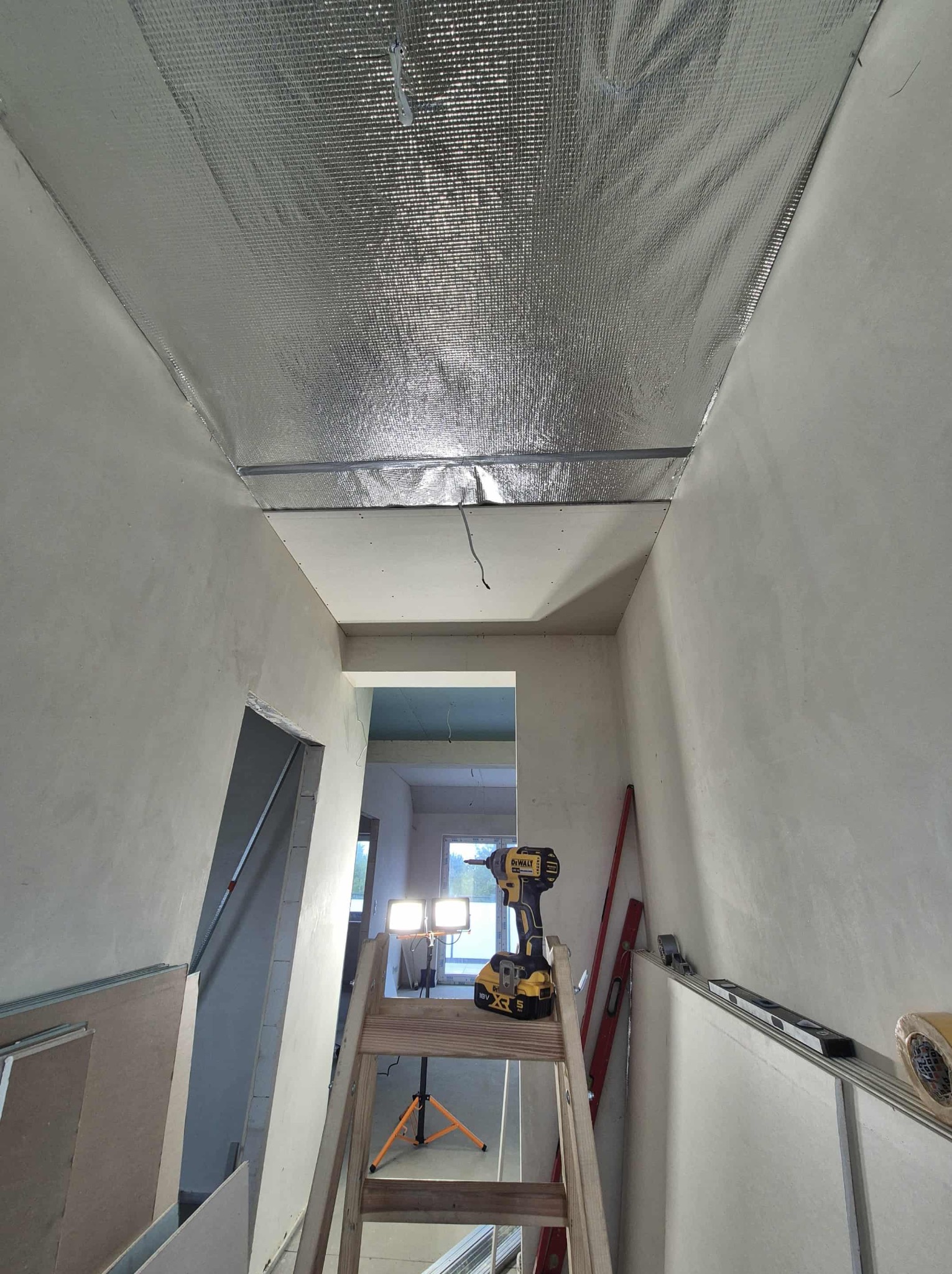 Montaż izolacji termicznej sufitu folią aluminiową w Pogórzu. Widoczna drabina, wkrętarka i poziomica. Prace wykończeniowe w trakcie realizacji.