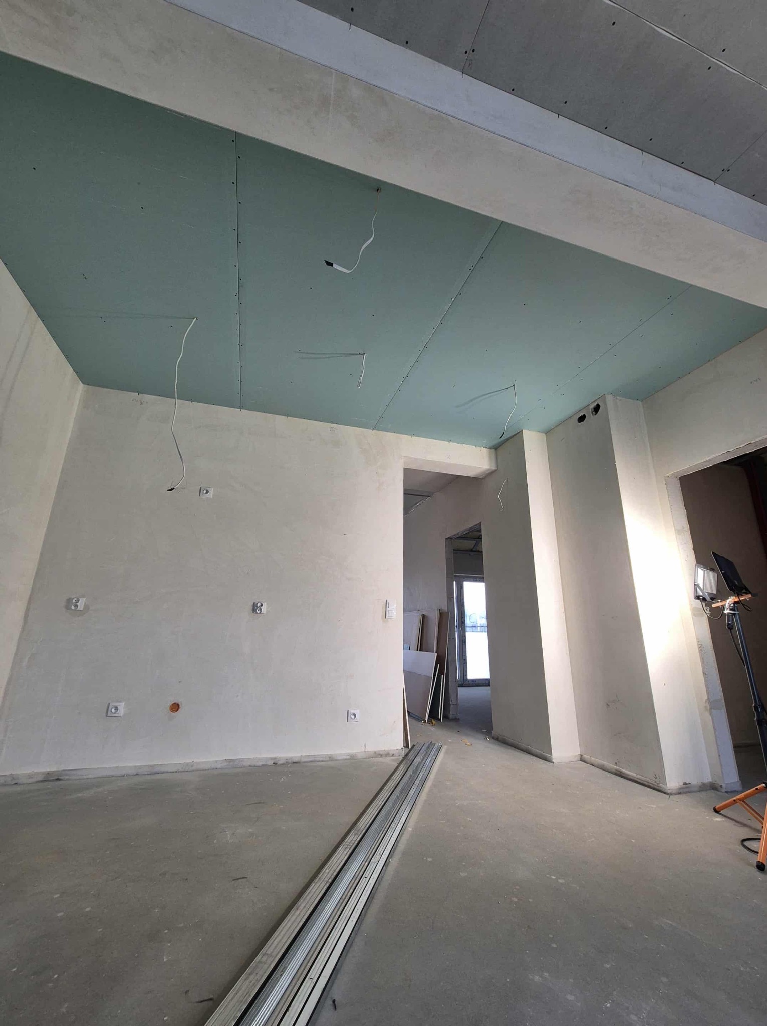 Wnętrze w trakcie prac wykończeniowych: sufit z płyt kartonowo-gipsowych, ściany po szpachlowaniu, widoczne profile na podłodze i lampa budowlana. Stan surowy.