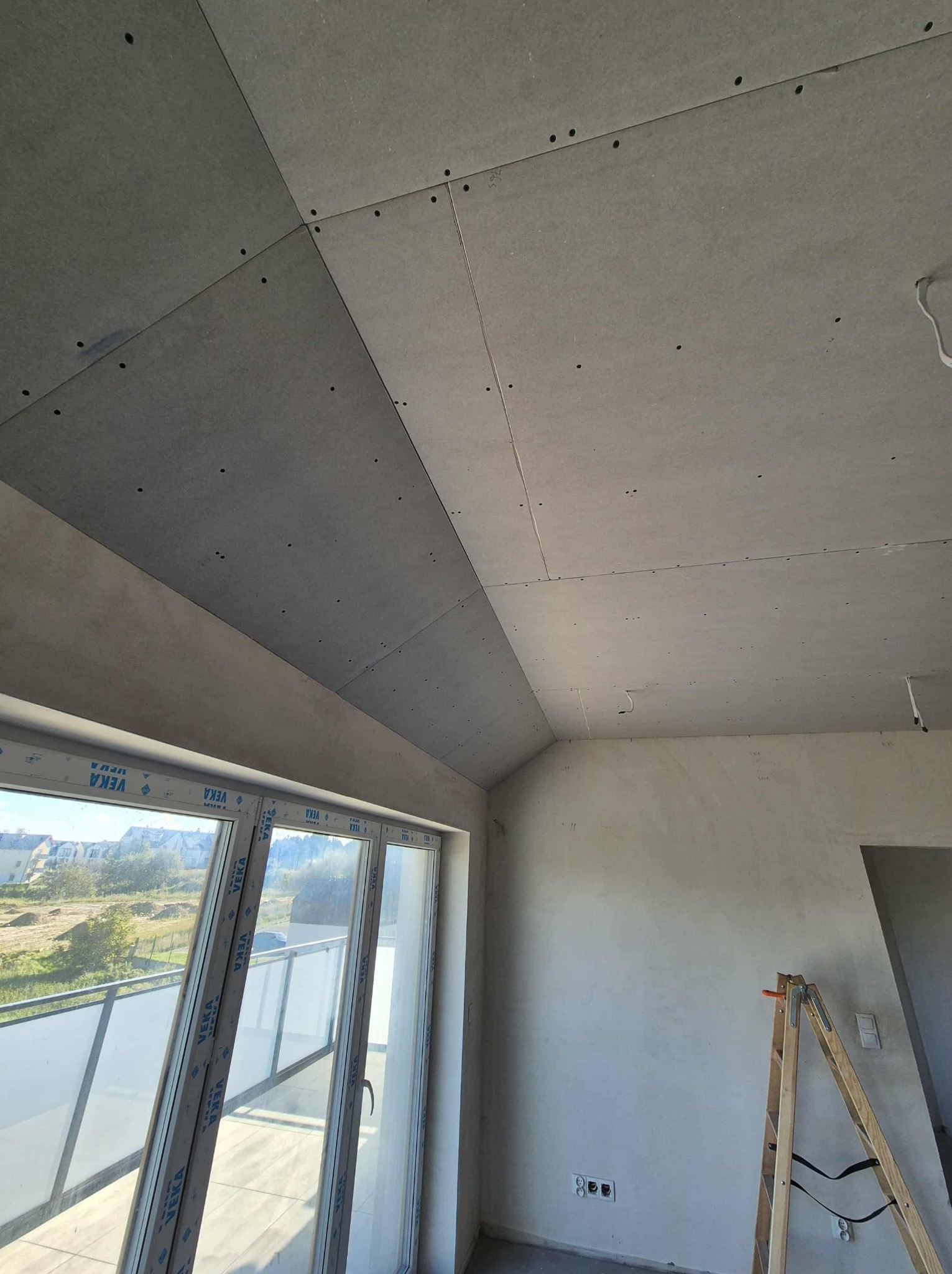 Skośny sufit z płyt gipsowo-kartonowych w trakcie montażu, widoczne łączenia i otwory na wkręty. W tle okno balkonowe i fragment ściany z drabiną.