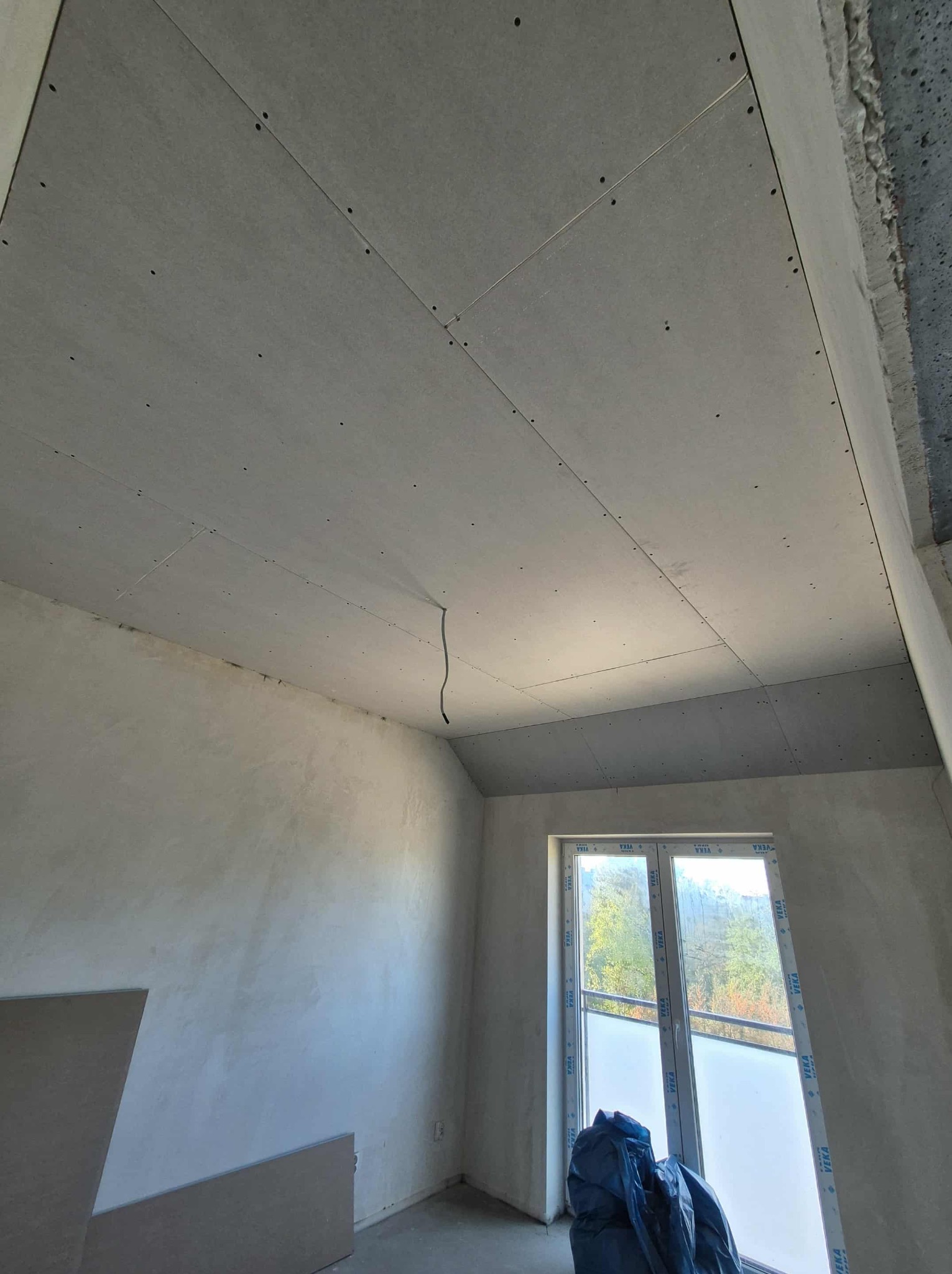 Pokój w trakcie prac wykończeniowych: zamontowane płyty na suficie, ściany po szpachlowaniu, okno z widokiem na zieleń. Widoczny kabel zwisający z sufitu.