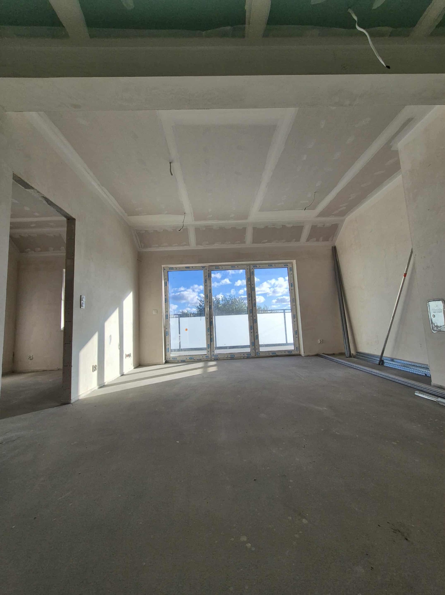 Surowe wnętrze z oknem balkonowym, ściany przygotowane pod malowanie, widoczne płyty gipsowe na suficie i profile metalowe. Jasne, przestronne pomieszczenie w trakcie prac wykończeniowych.