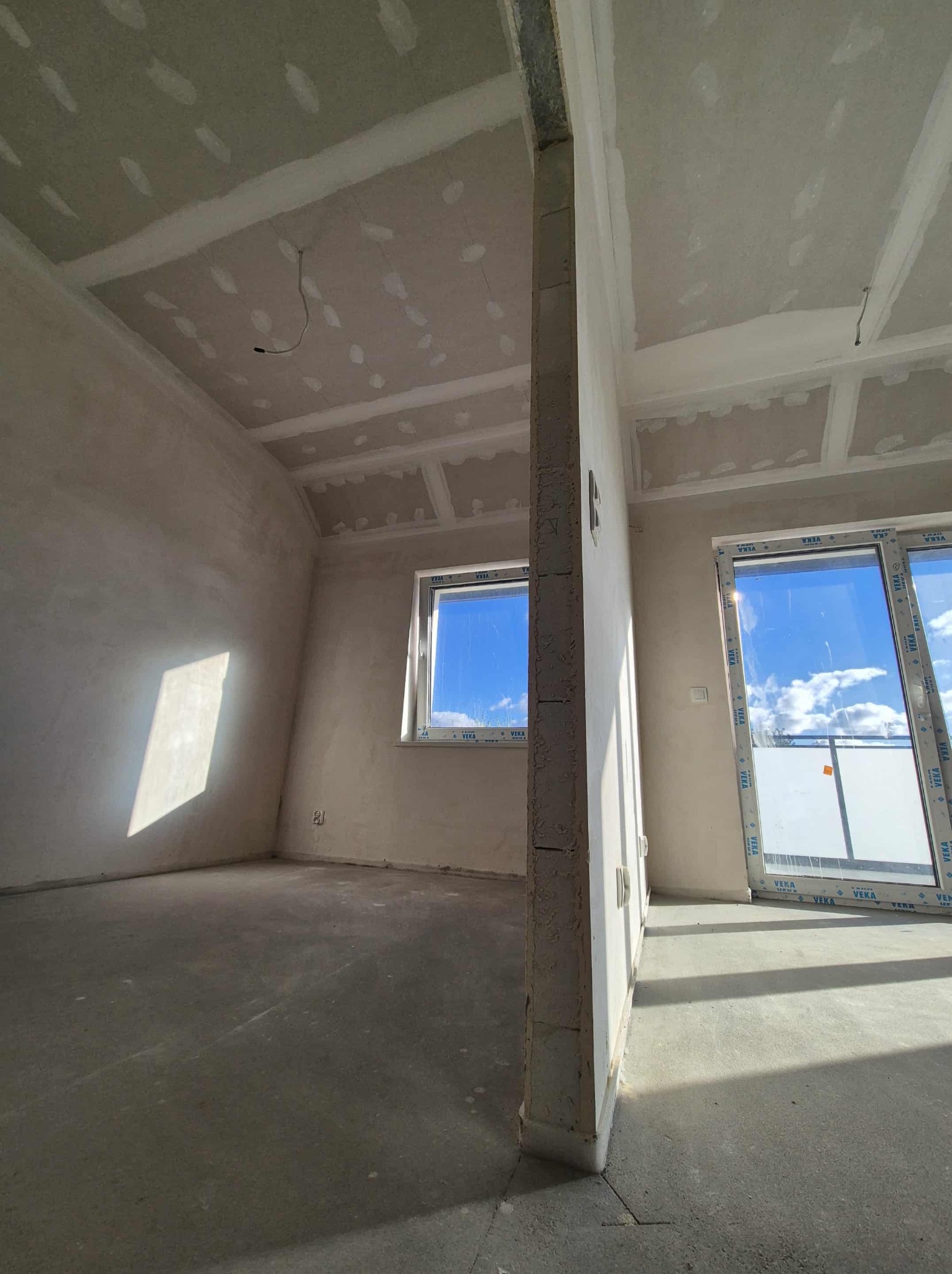 Wnętrze w trakcie wykończenia: surowy beton na podłodze, ściany po szpachlowaniu, płyty karton-gips na suficie z widocznymi punktami szpachlowania. Okno i drzwi balkonowe z PCV.