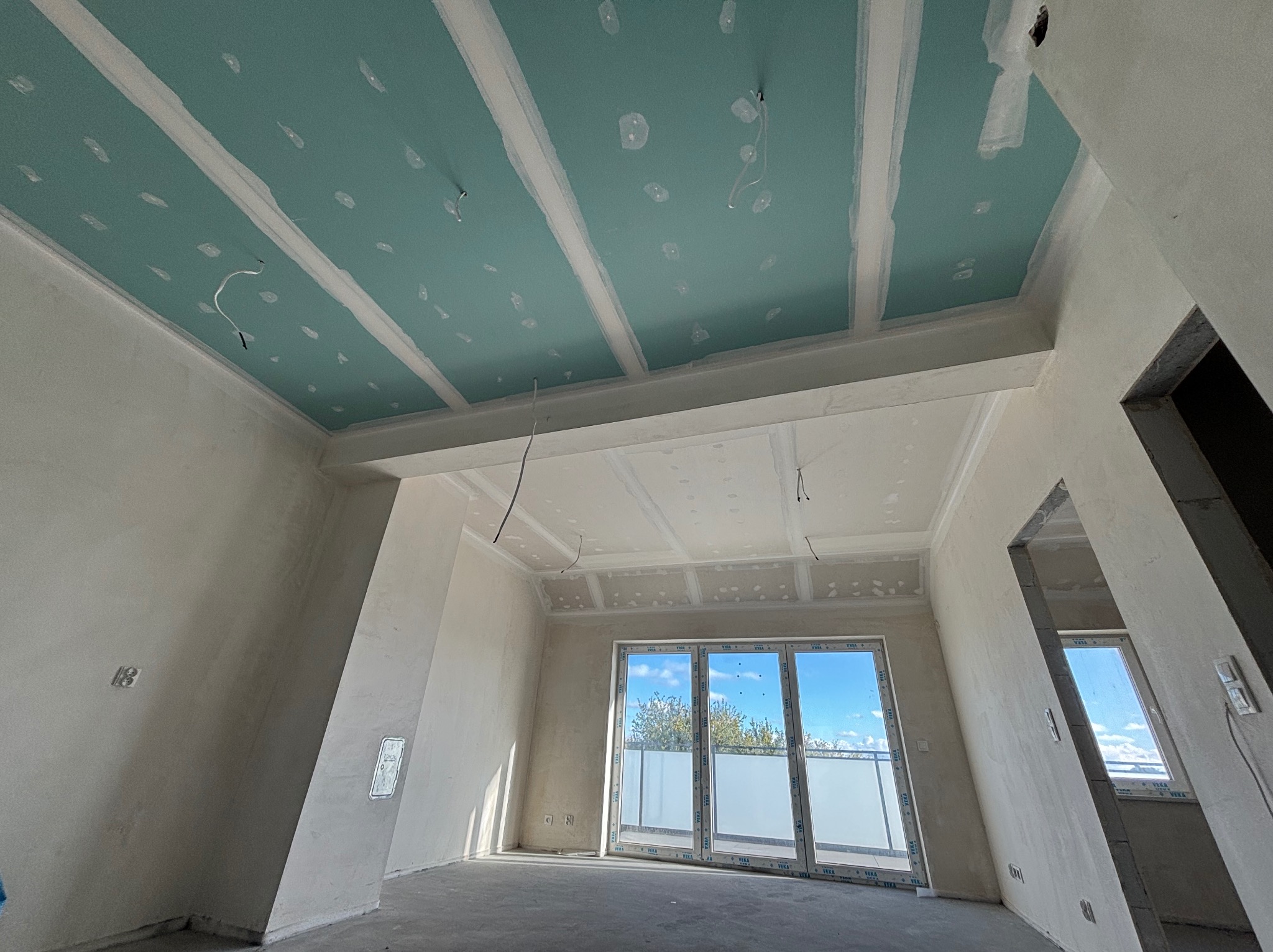 Wnętrze w trakcie prac: sufit z płyt gipsowo-kartonowych, wstępnie wyszpachlowany, widoczne przewody elektryczne. Ściany przygotowane do dalszych prac wykończeniowych.