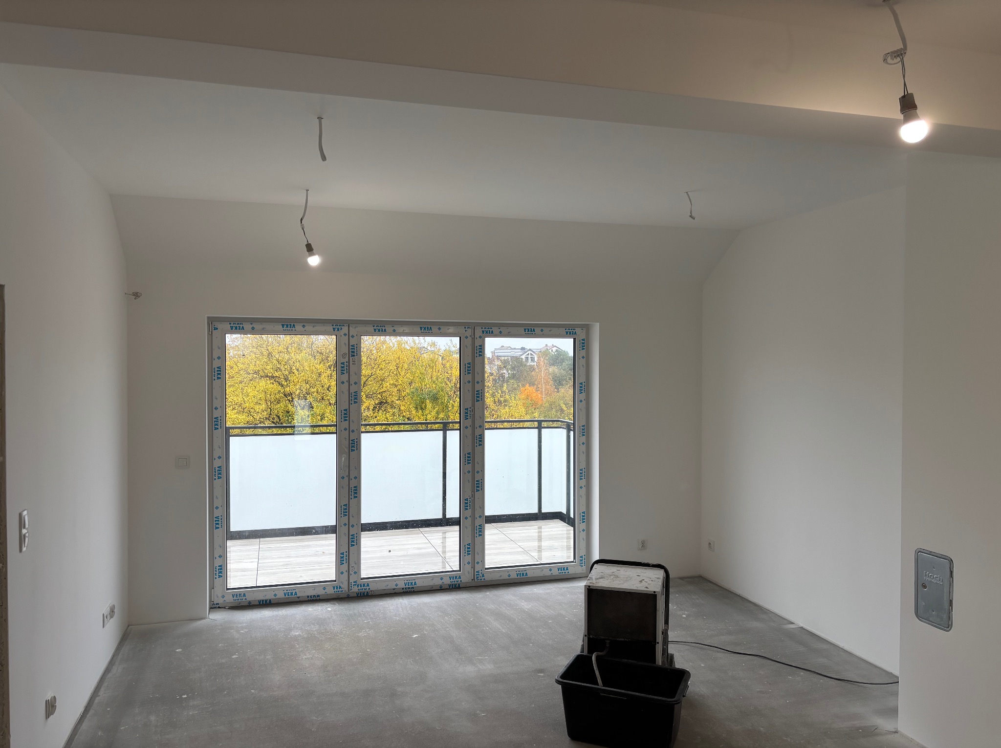 Białe, świeżo pomalowane wnętrze z dużym oknem balkonowym i widokiem na jesienne drzewa. Na podłodze stoi urządzenie budowlane. Minimalistyczny styl wykończenia.