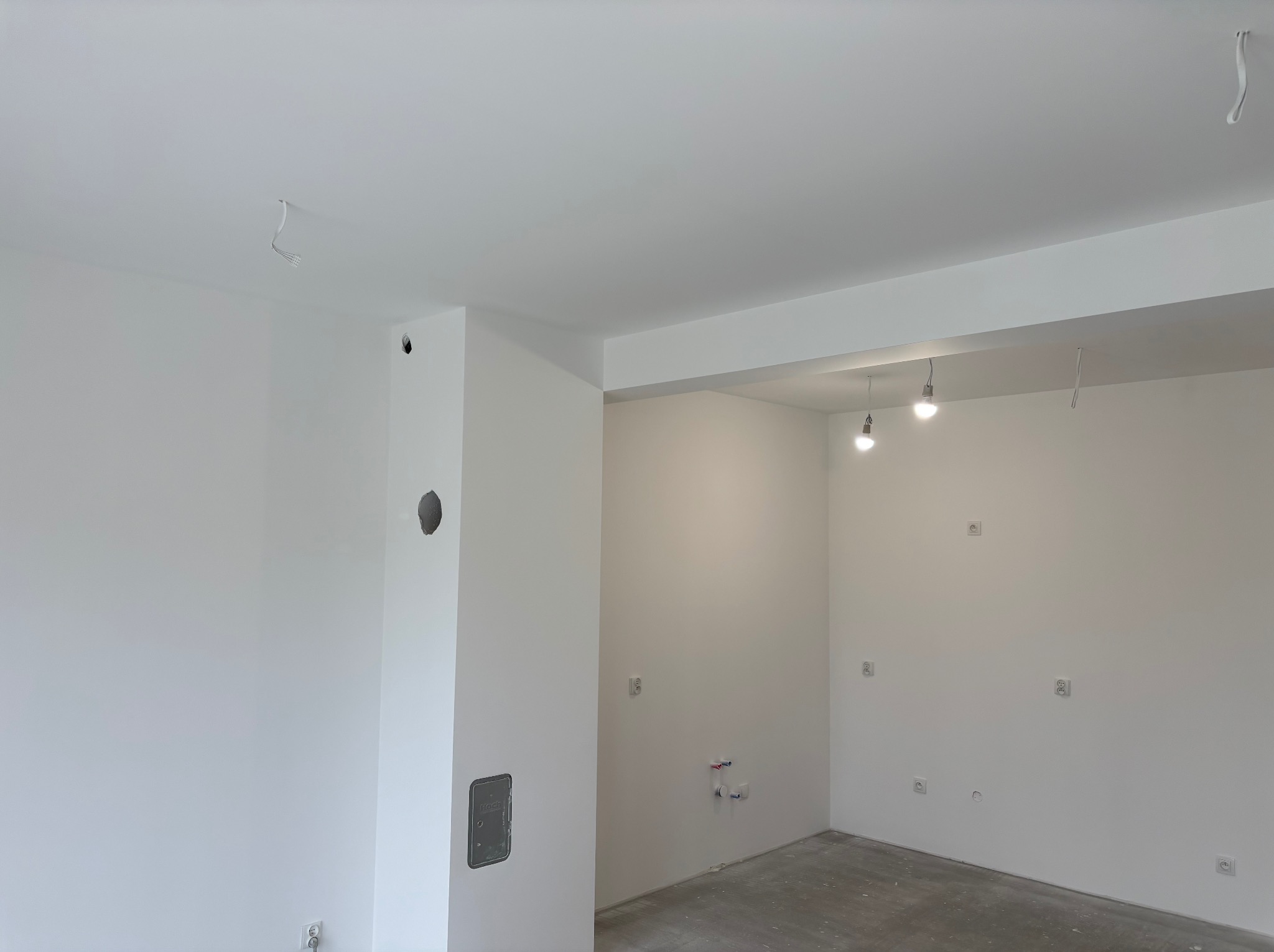 Białe, świeżo pomalowane wnętrze z widocznymi gniazdkami elektrycznymi i wyprowadzonymi kablami oświetleniowymi. Minimalistyczny design, surowy beton na podłodze.