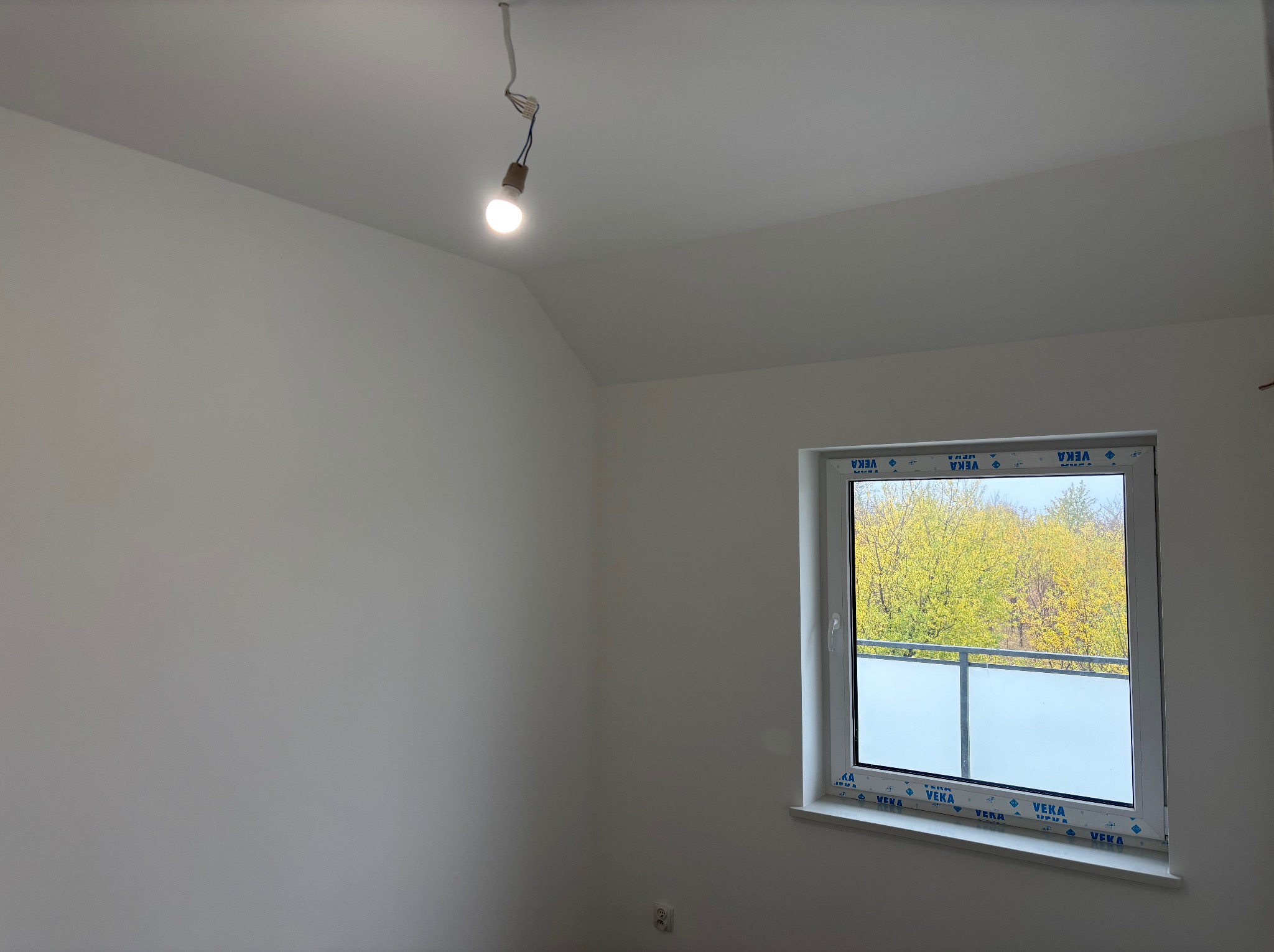 Wnętrze pokoju po remoncie: białe ściany, okno z widokiem na drzewa, surowa żarówka. Minimalistyczne wykończenie, jasne, naturalne światło.