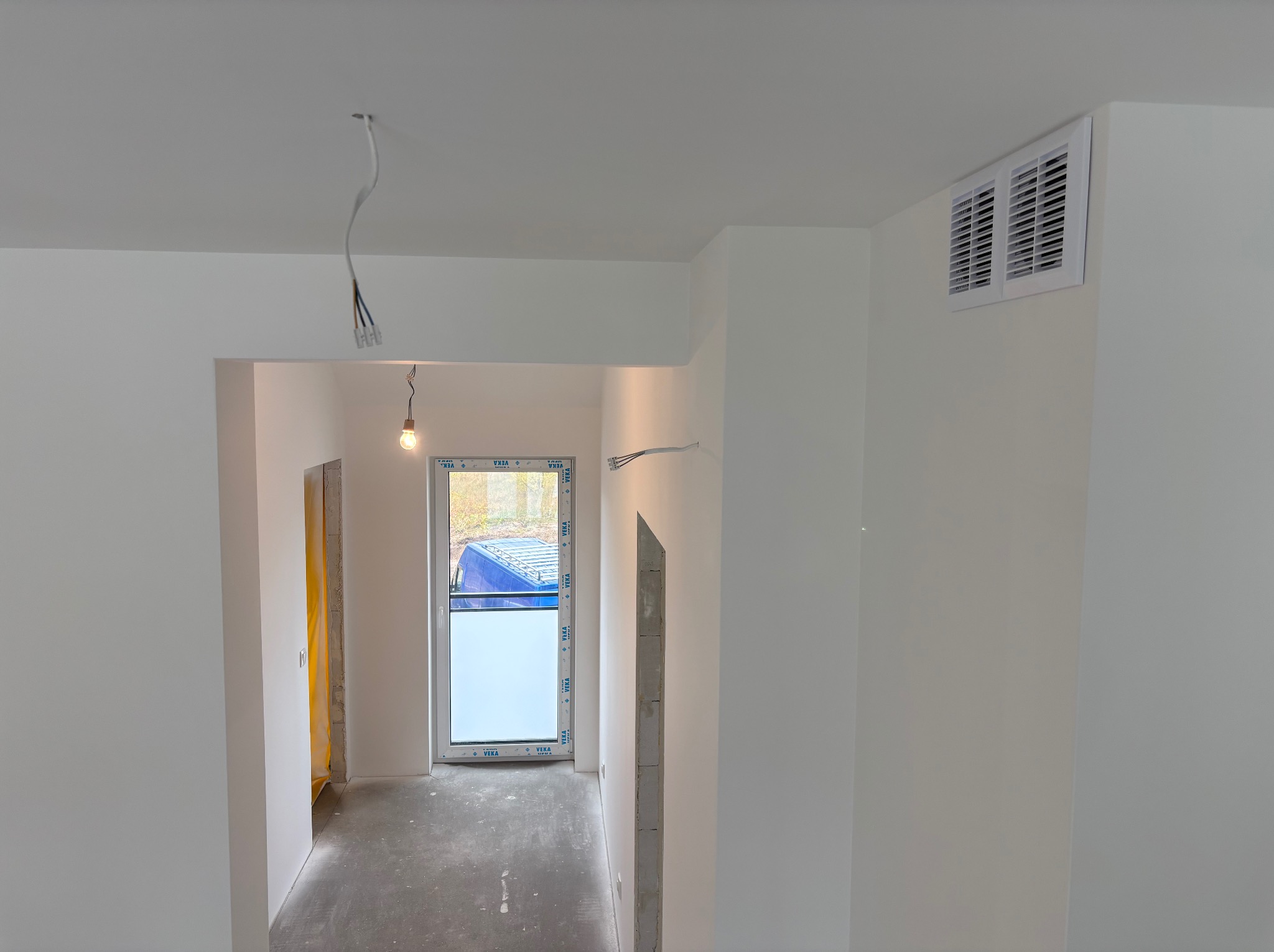 Białe, świeżo pomalowane wnętrze z widoczną kratką wentylacyjną, oknem z naklejkami i wiszącymi kablami, ukazujące etap wykończenia.