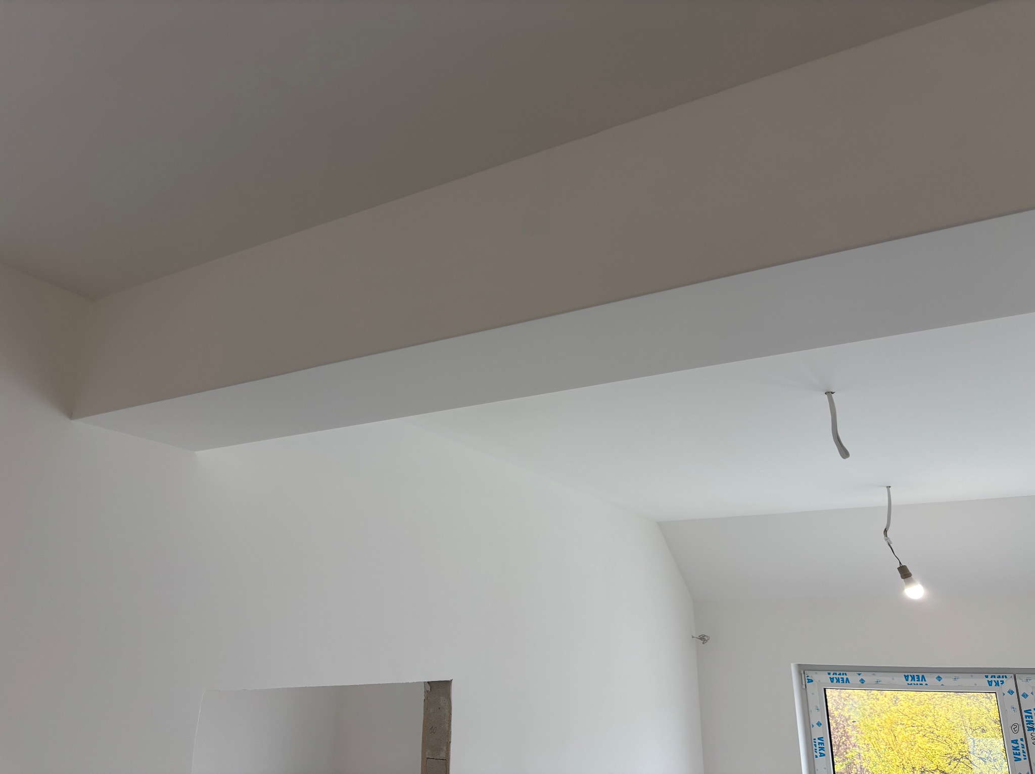 Białe, świeżo pomalowane wnętrze z widoczną konstrukcją sufitu podwieszanego i oknem z widokiem na zieleń. Minimalistyczny design, surowy stan wykończenia.