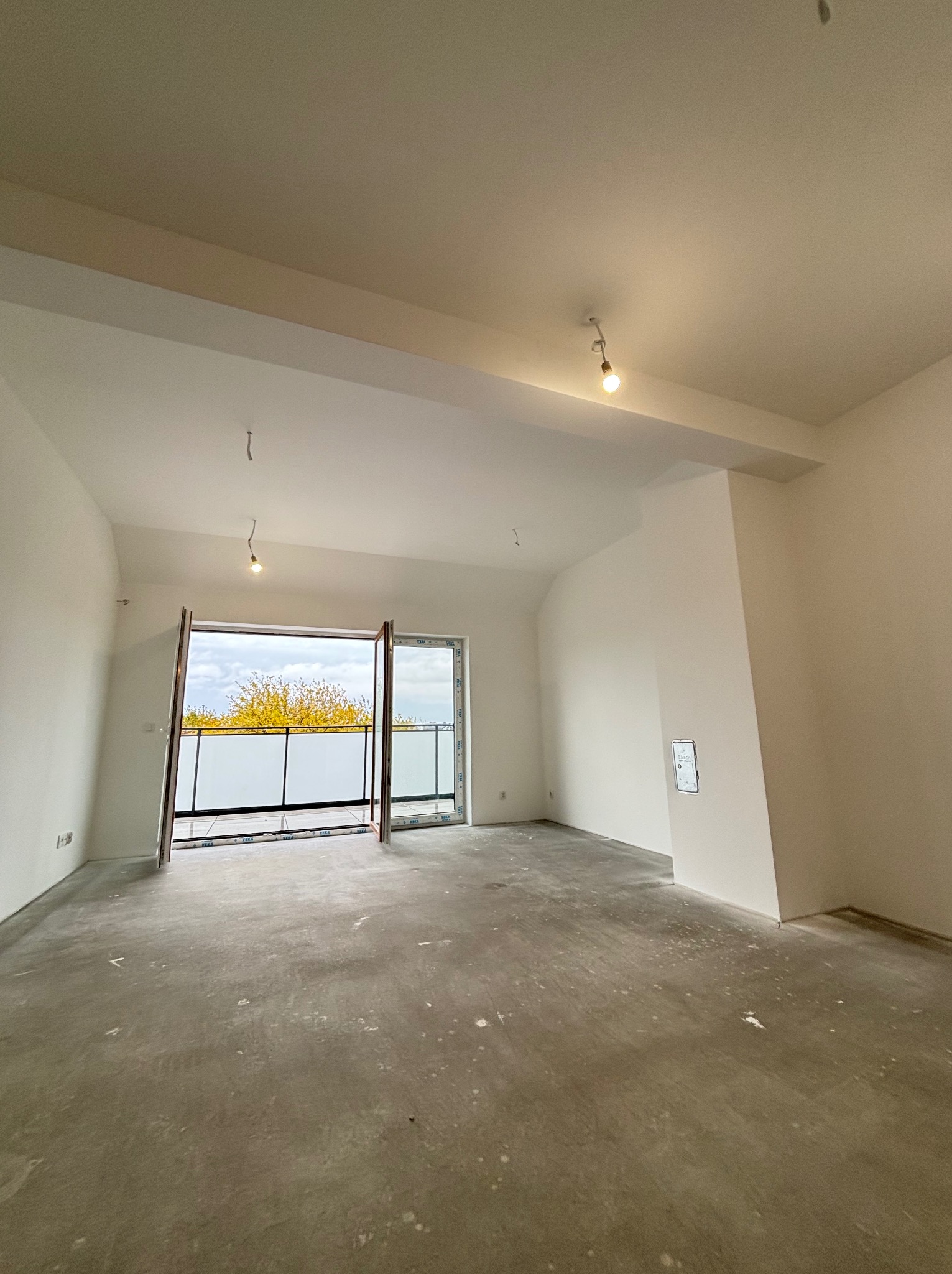 Jasne, puste wnętrze z otwartymi drzwiami balkonowymi, surowy beton na podłodze, świeżo pomalowane ściany na biało, widok na zewnątrz z żółtymi drzewami.