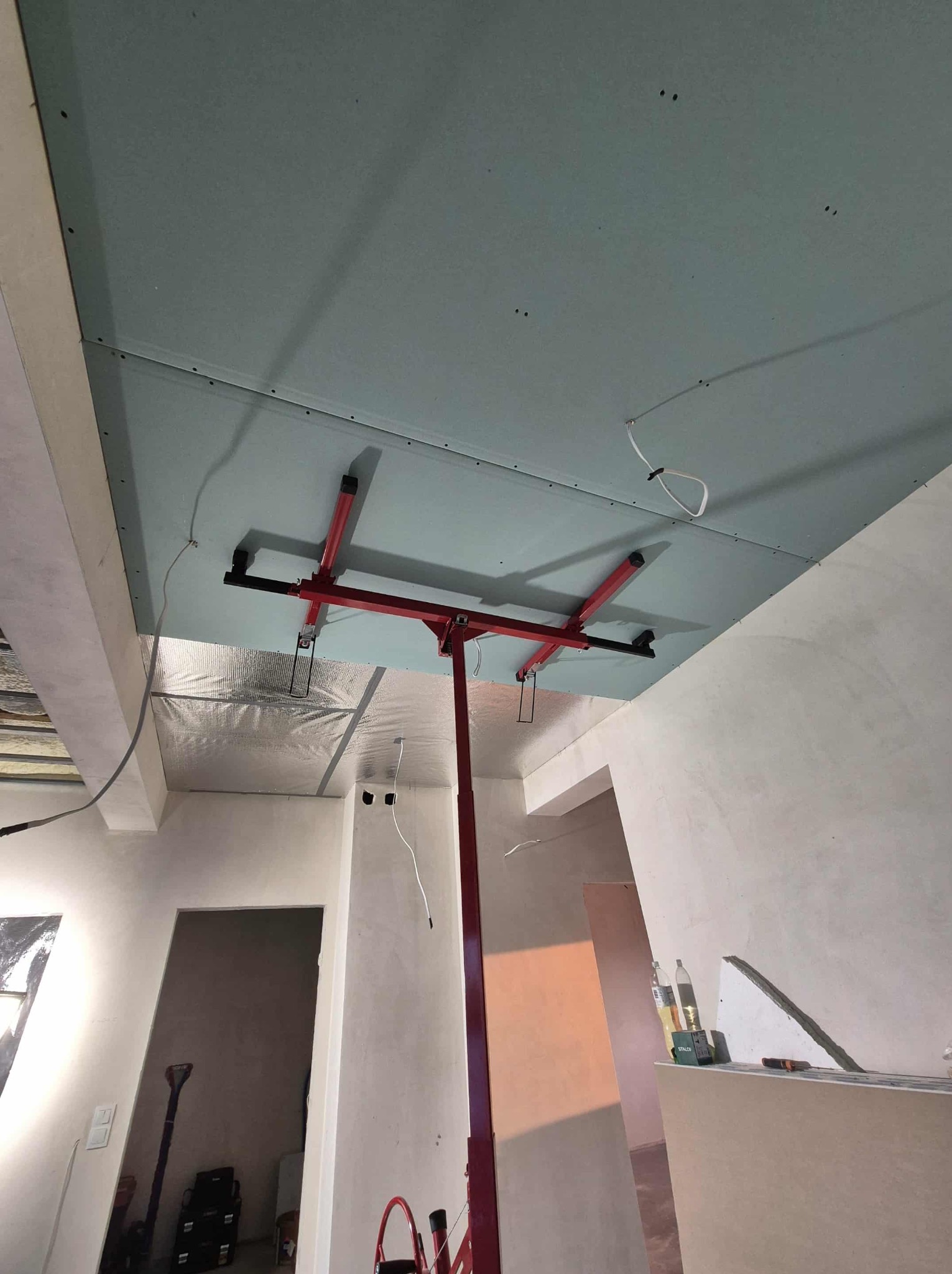 Montaż płyty gipsowej na suficie za pomocą podnośnika. Widoczne łączenia płyt i częściowo wykończone ściany. W tle folia izolacyjna i surowy stan pomieszczenia.
