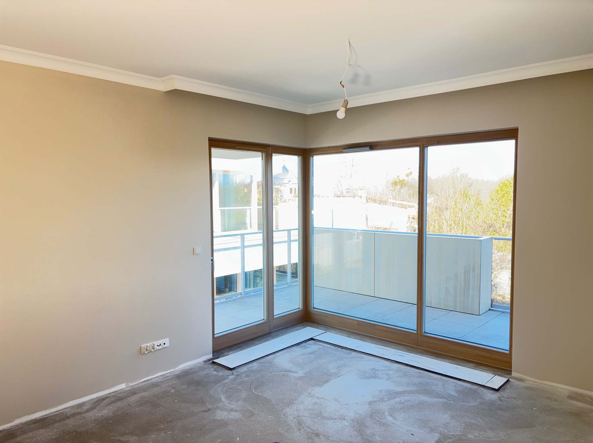 Jasne wnętrze z beżowymi ścianami, białym sufitem z listwami i dużymi oknami balkonowymi. Na podłodze widoczne płyty przygotowane do montażu.