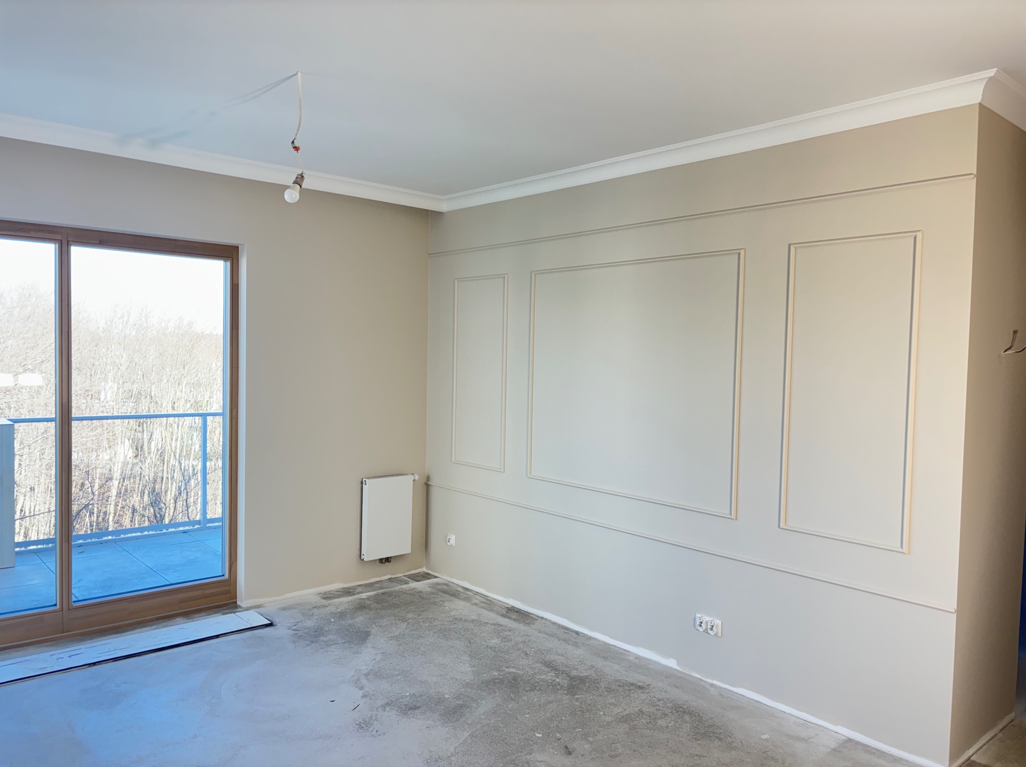 Jasne wnętrze po remoncie z dekoracyjnymi panelami ściennymi i listwami przysufitowymi. Widoczne duże okno balkonowe i surowa podłoga przed wykończeniem.